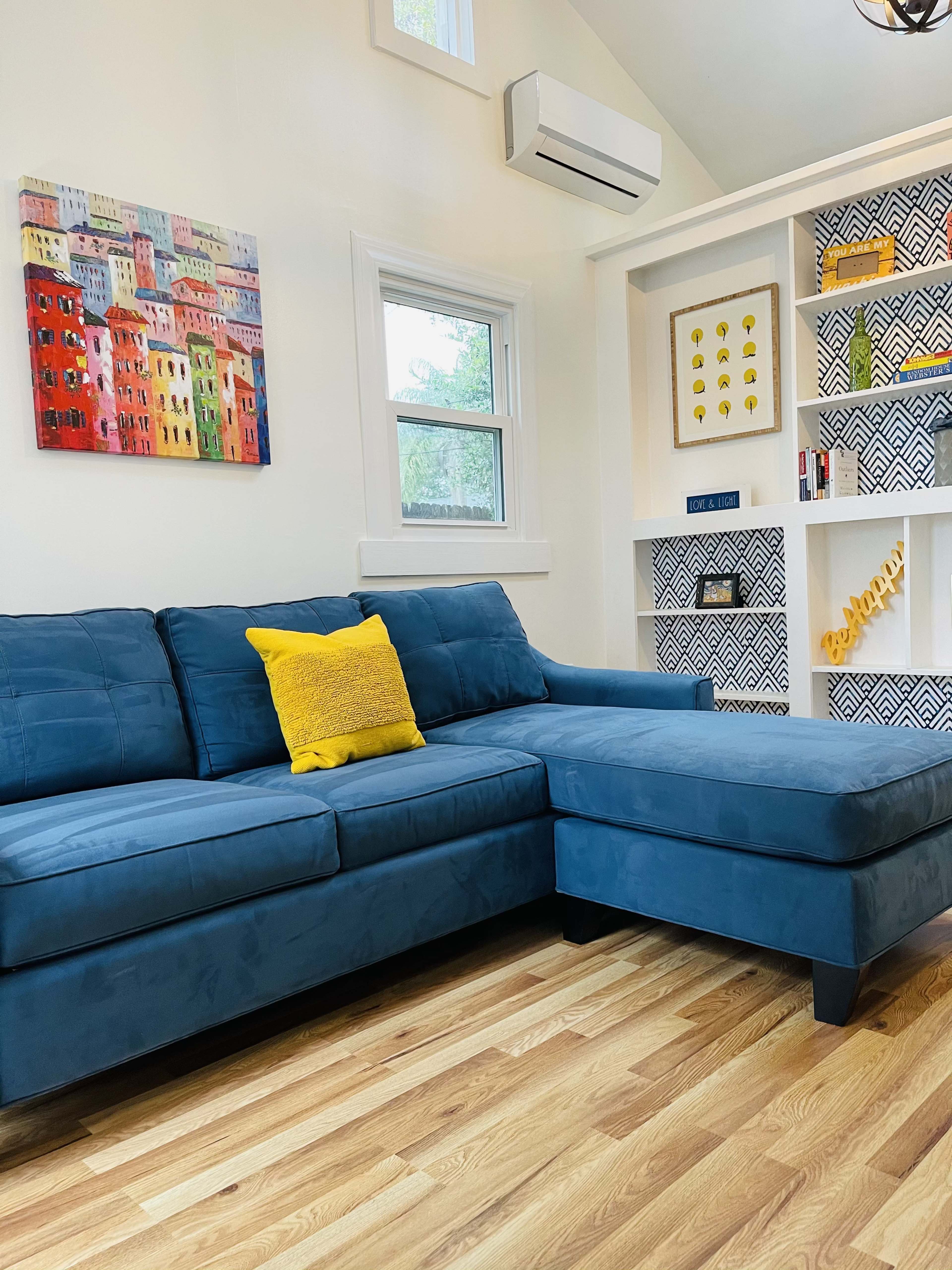 A blue sectional sofa with a yellow pillow is positioned in a bright room featuring a colorful wall art piece and patterned shelves.