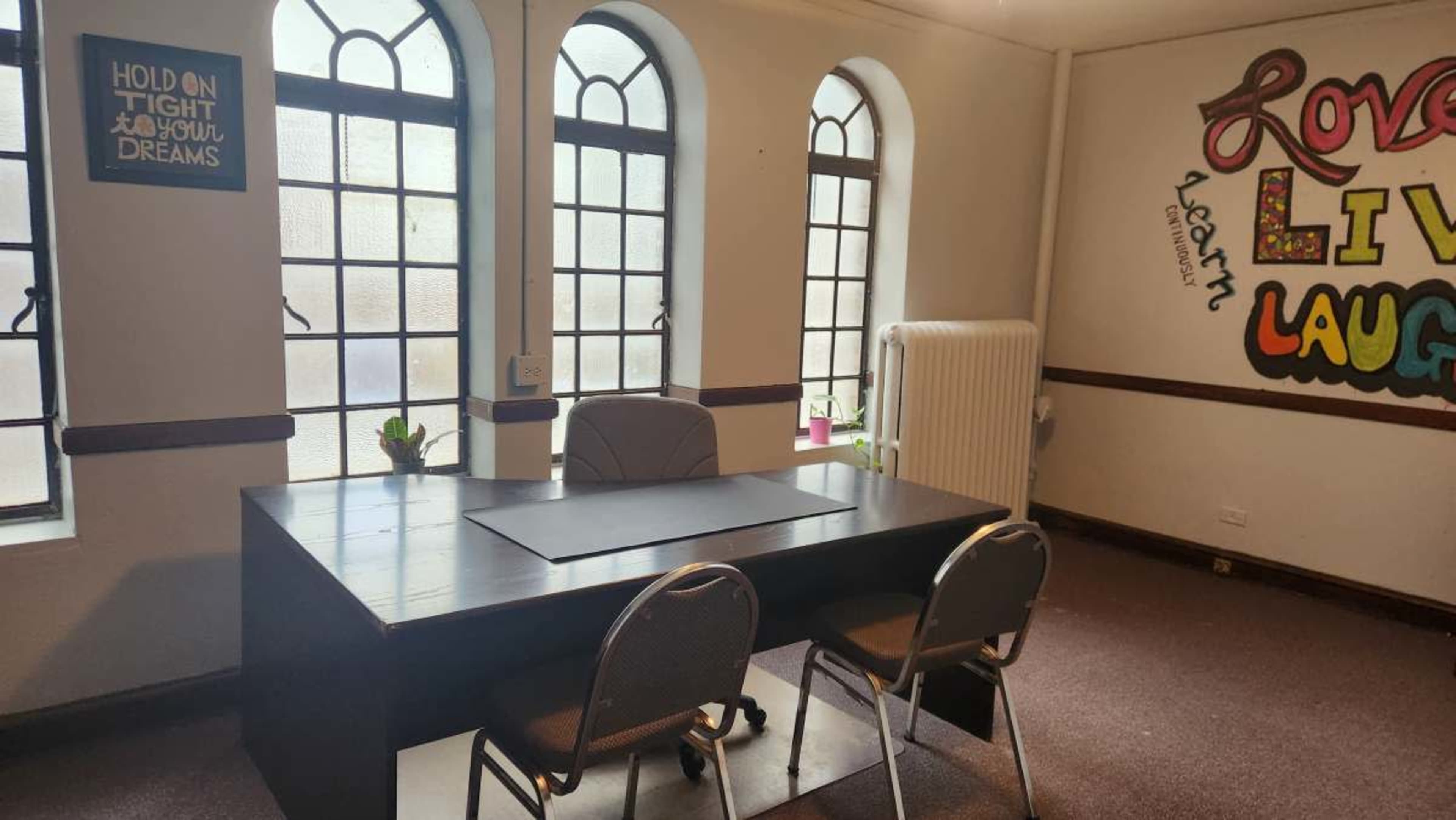 The image shows a simple office space featuring a black desk with two chairs, large arched windows, and a colorful wall mural that includes the words "Love," "Live," and "Laugh."