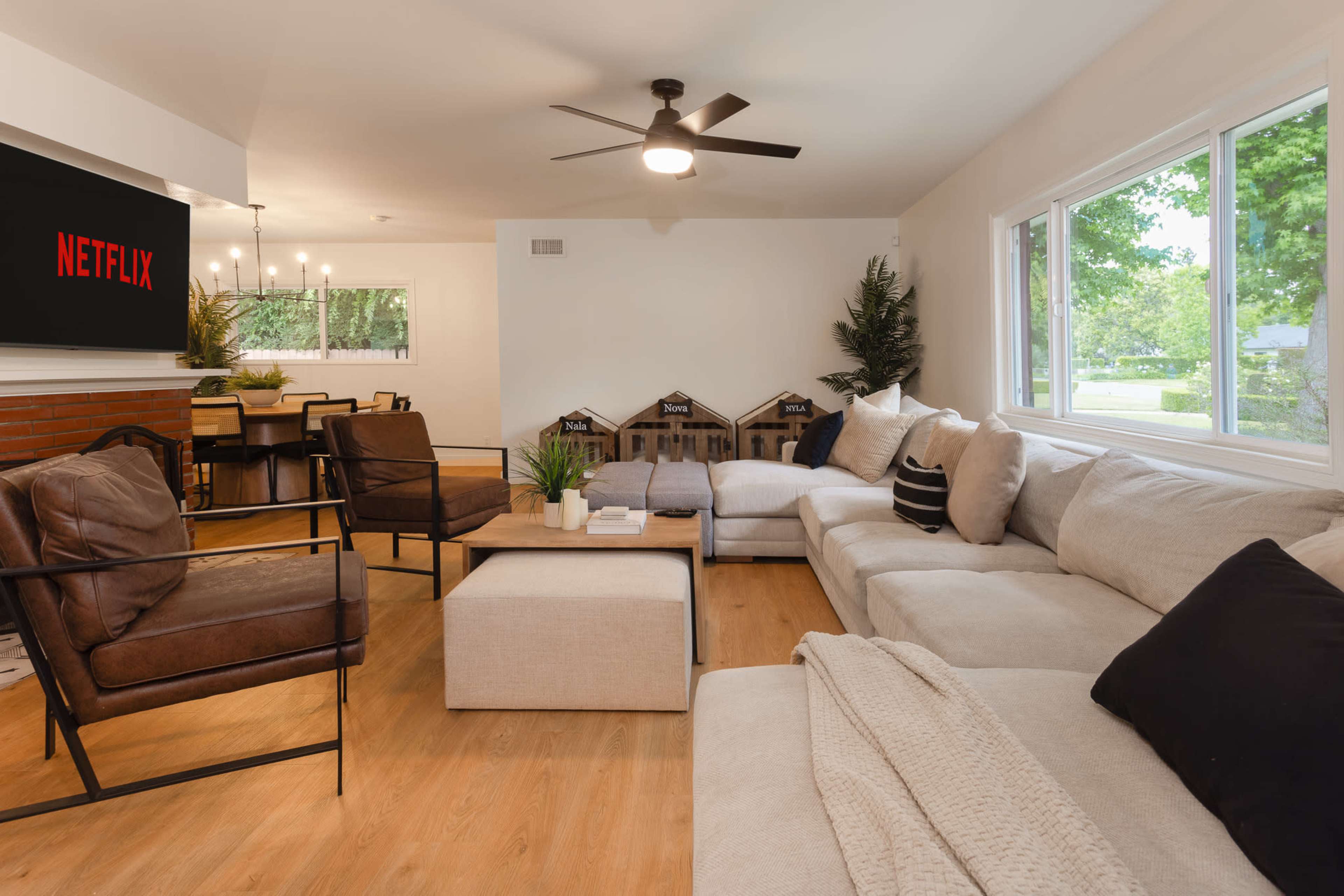 A spacious living room with a large sectional sofa, a coffee table, and a wall-mounted TV displaying the Netflix logo, all illuminated by natural light from a nearby window.