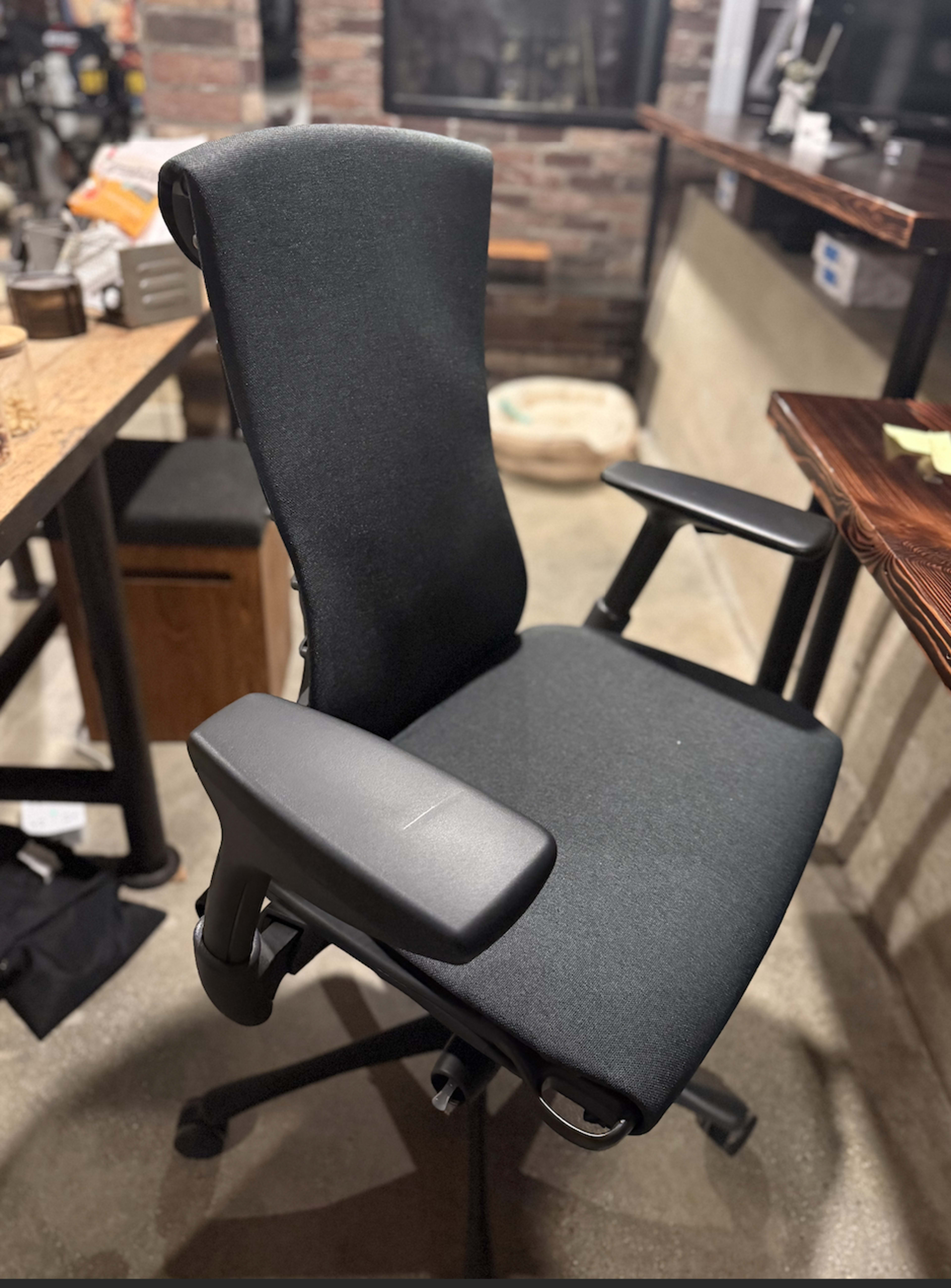 The image shows a black ergonomic office chair with adjustable armrests, positioned in an indoor space with a rustic backdrop.