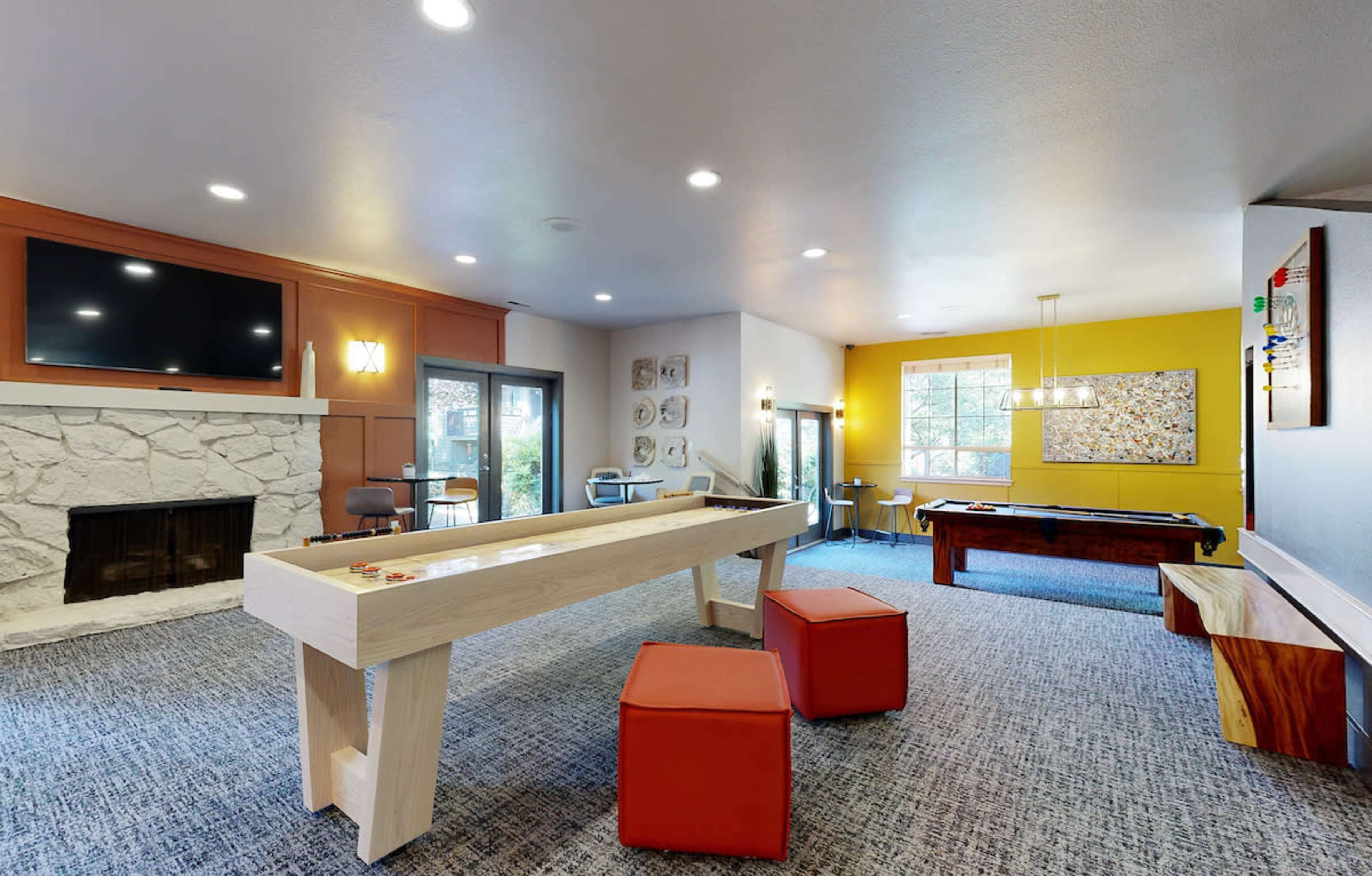 A modern recreational room featuring a fireplace, a large flat-screen TV, a shuffleboard table, and a billiards table, with colorful accents and seating areas.