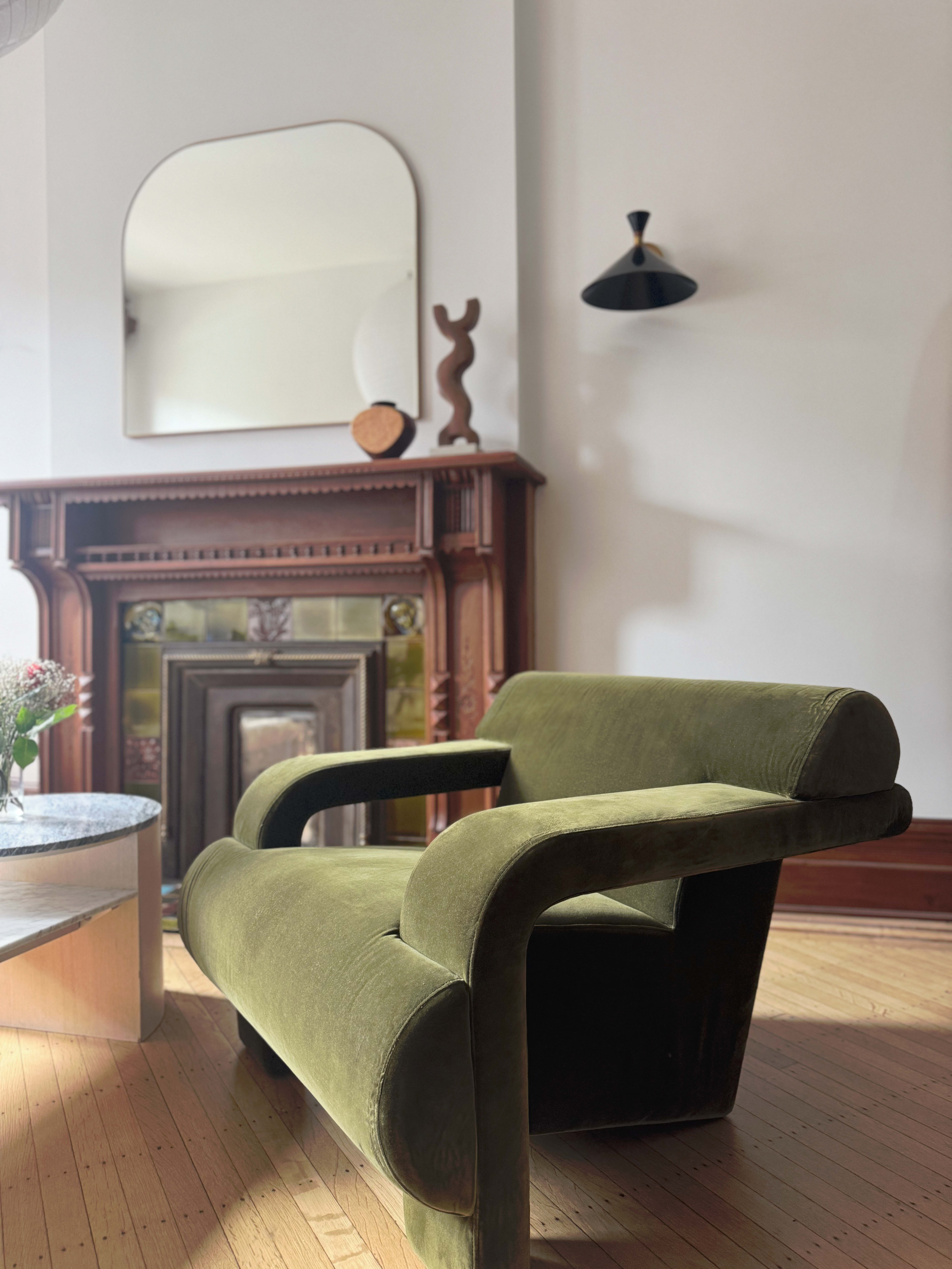 A green upholstered chair is positioned in a room featuring a wooden fireplace, a round table, and a mirror on the wall.