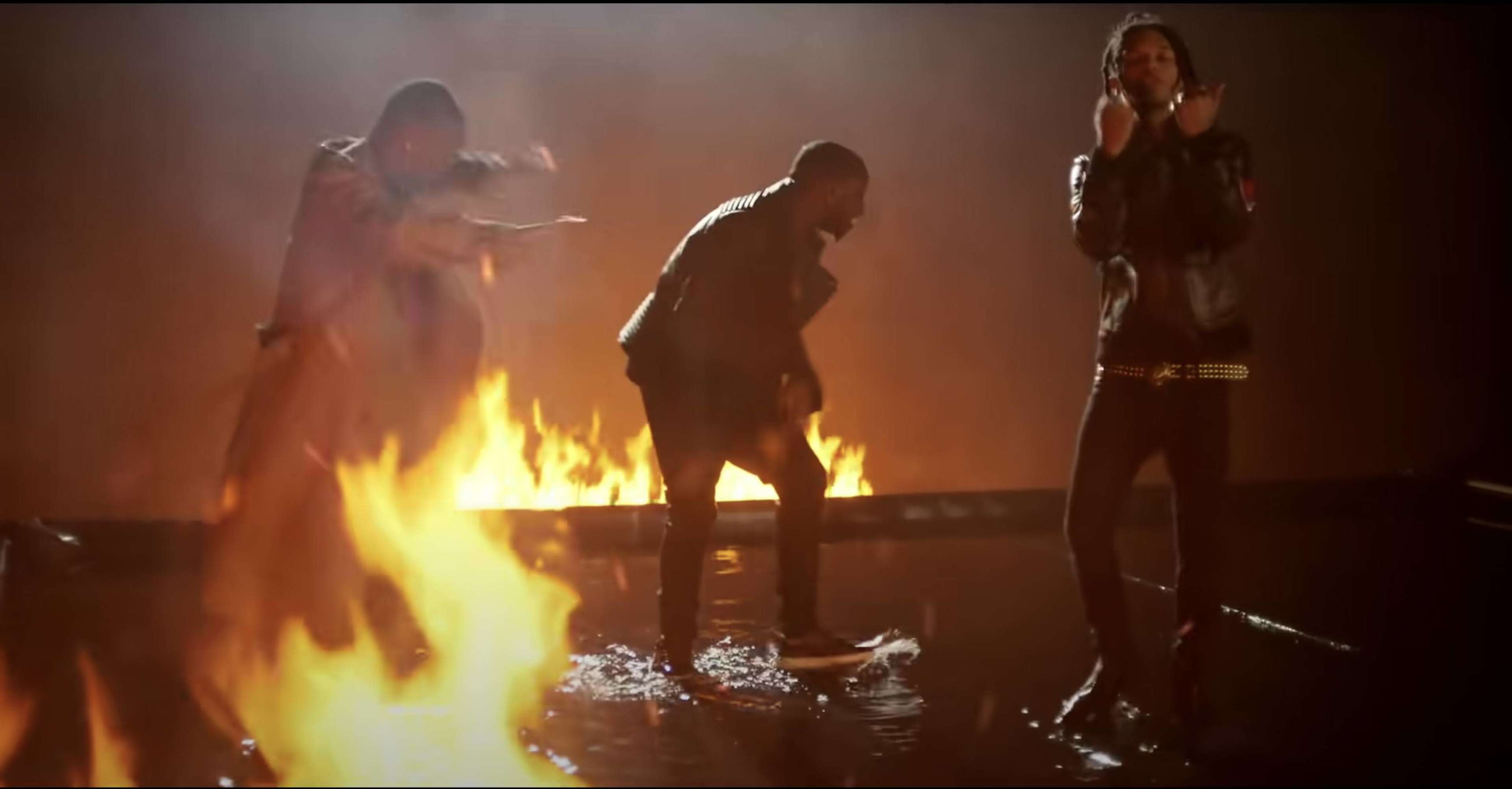 Three individuals stand on a wet surface amidst flames in a darkened environment.