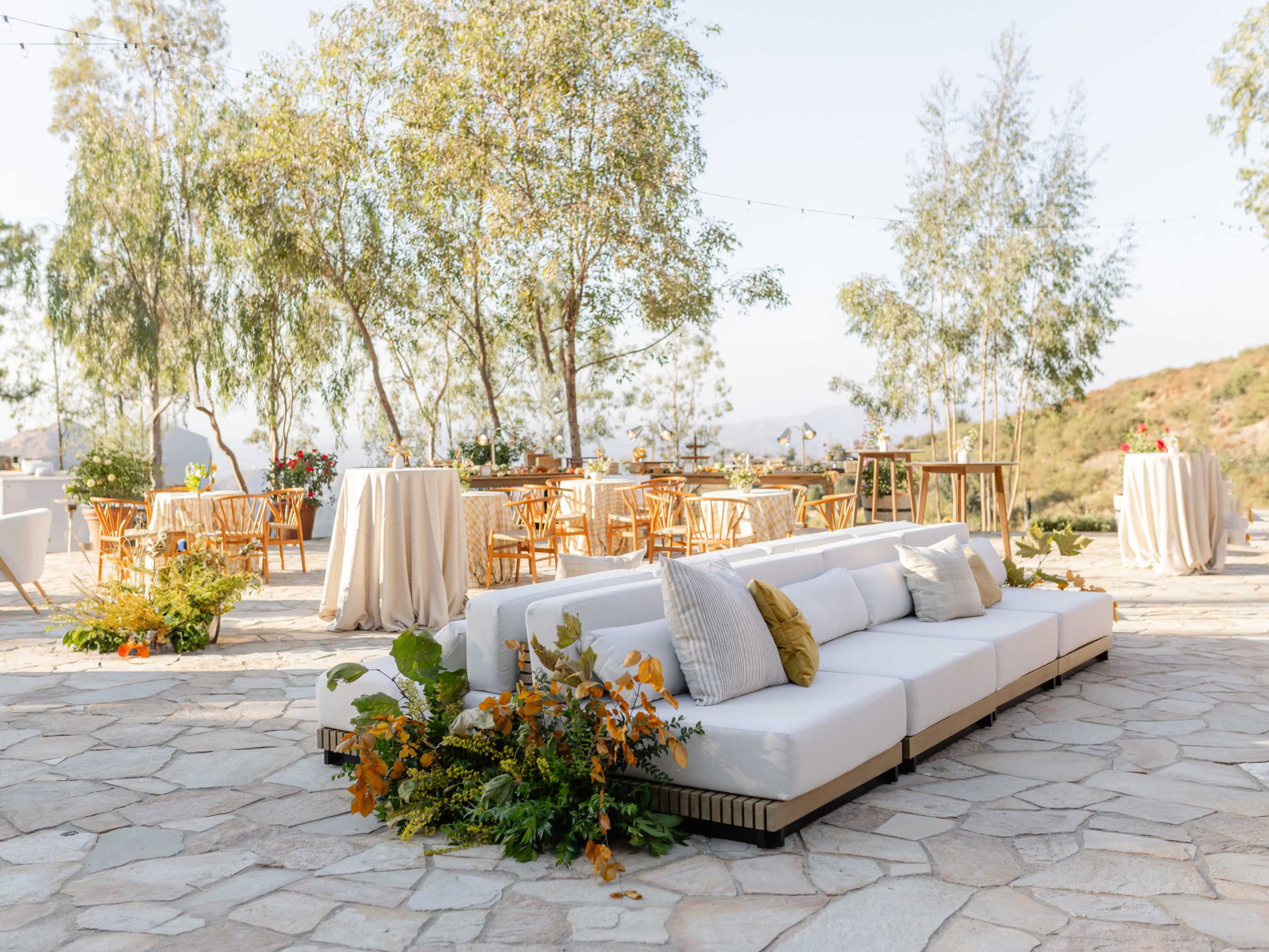 A modern outdoor lounge area features a white sectional sofa adorned with plants, surrounded by elegantly arranged tables and chairs beneath tall trees.