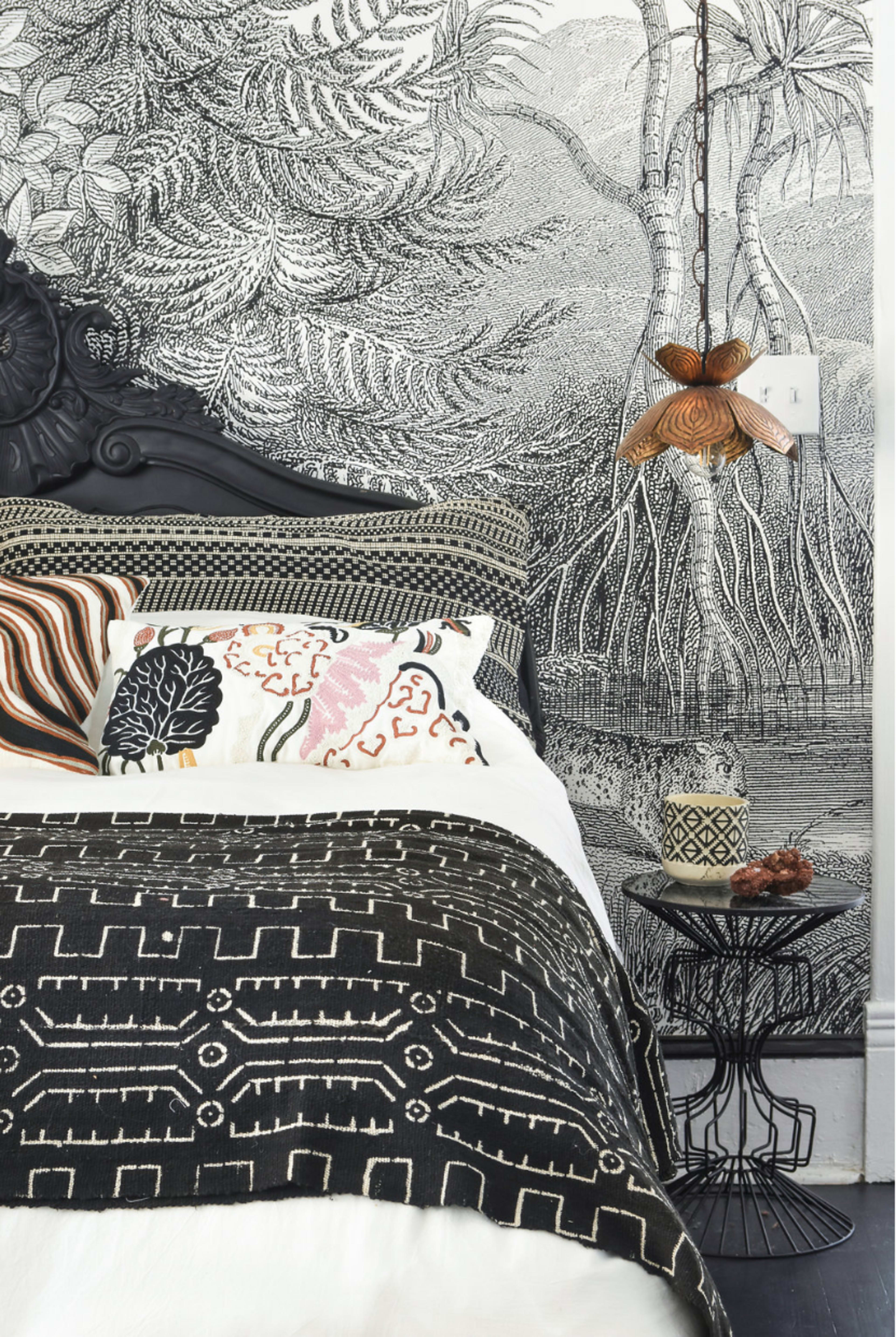 The image showcases a neatly made bed with patterned cushions and a decorative black-and-white wall mural featuring tropical foliage.