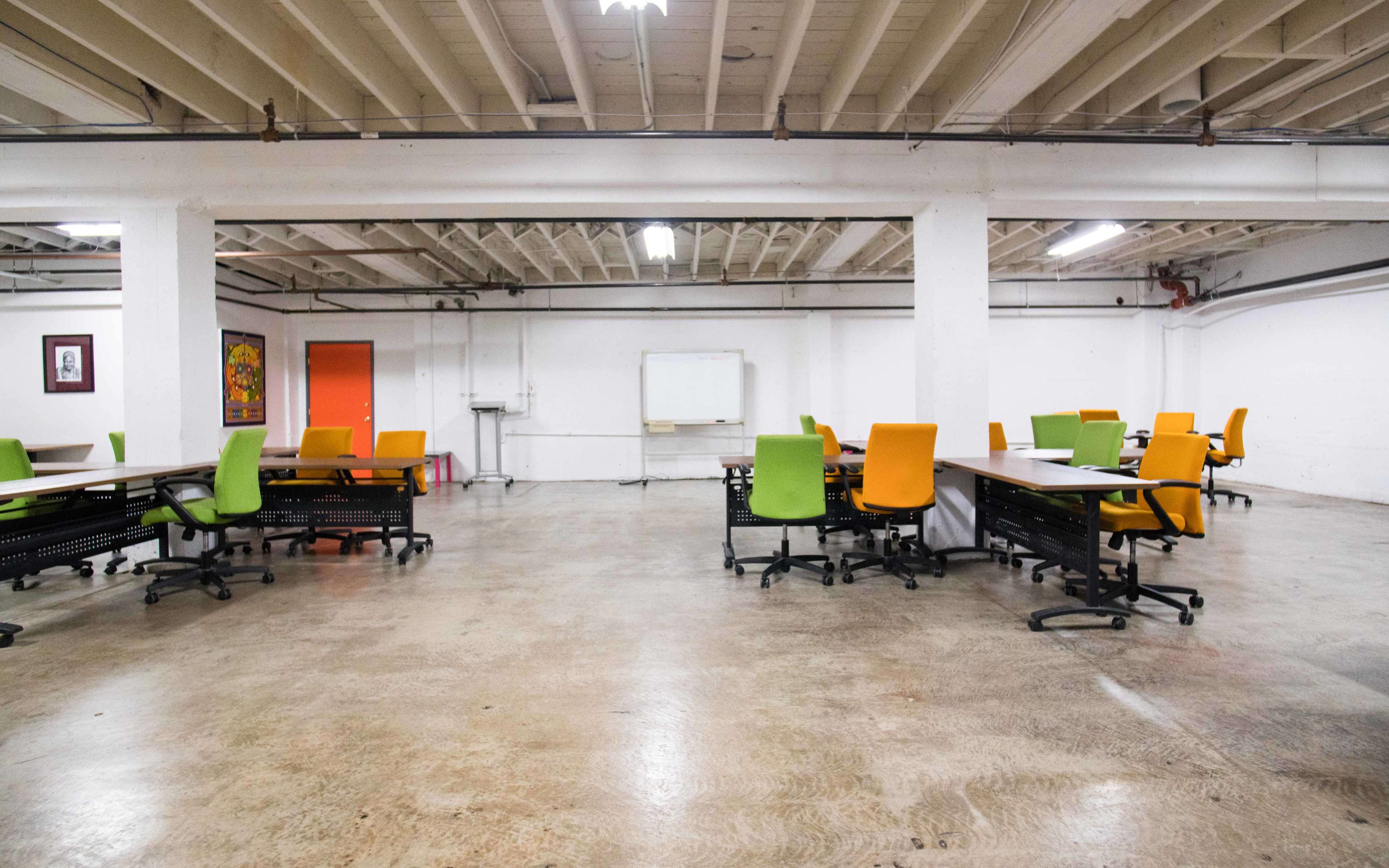 A spacious, empty office area featuring several desks with bright green and orange chairs, along with a whiteboard and orange door in the background.