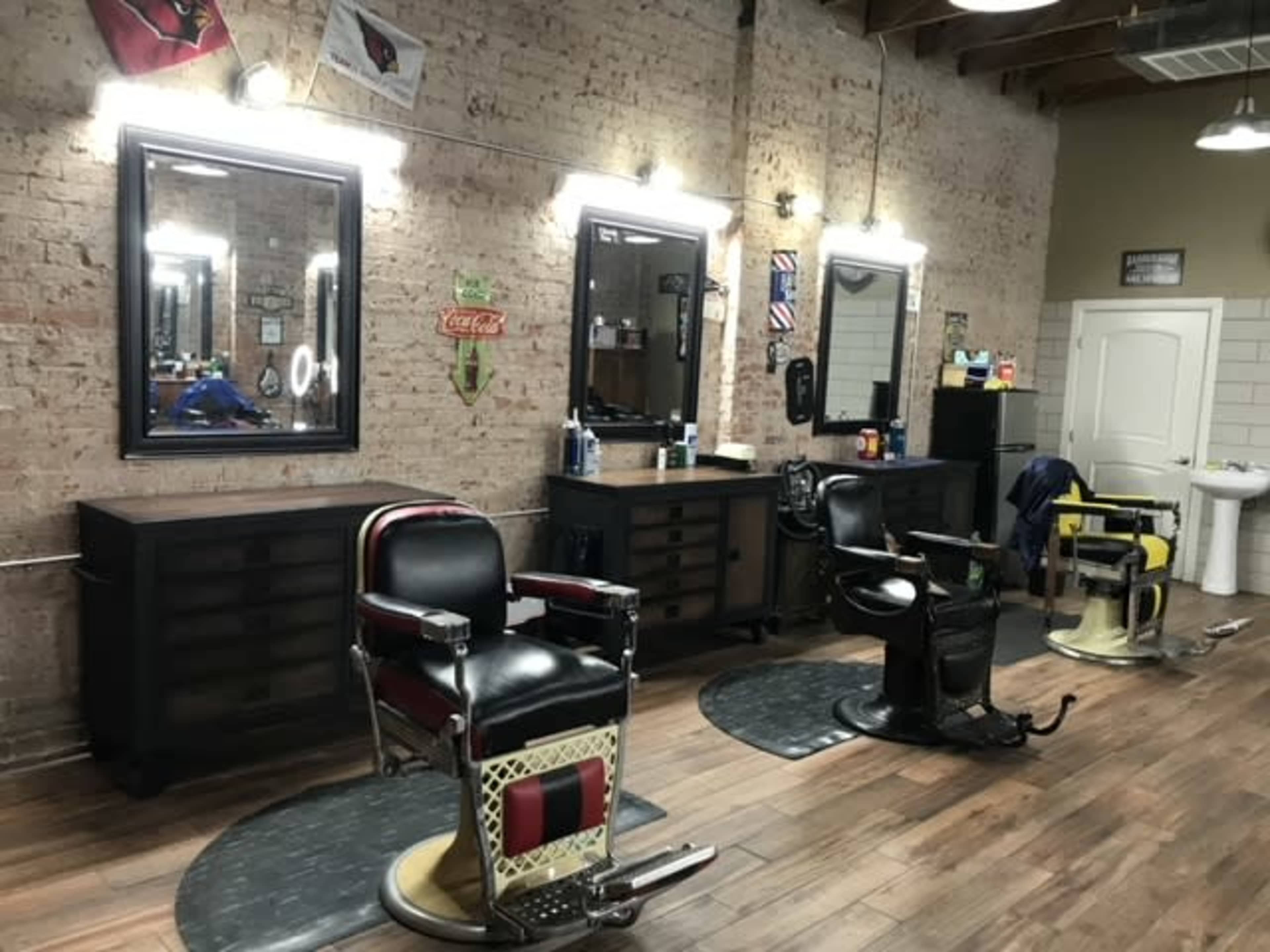 A barbershop interior with three vintage-style barber chairs and two large mirrors mounted on the wall, set against exposed brick and wooden floors.