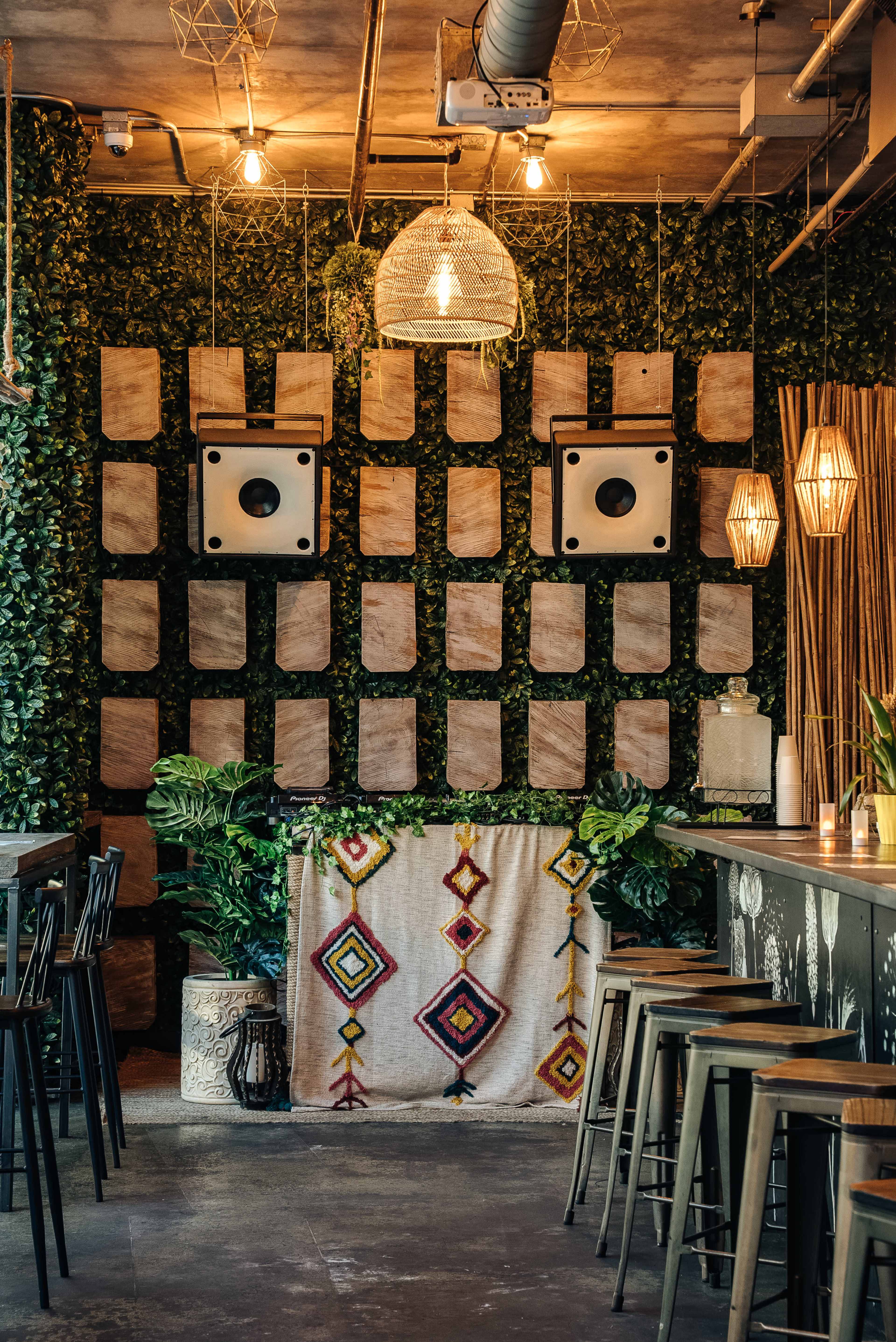 The image shows a bar area with a wall of green foliage, decorative wooden panels, and pendant lights, complemented by a woven textile draped over the counter.