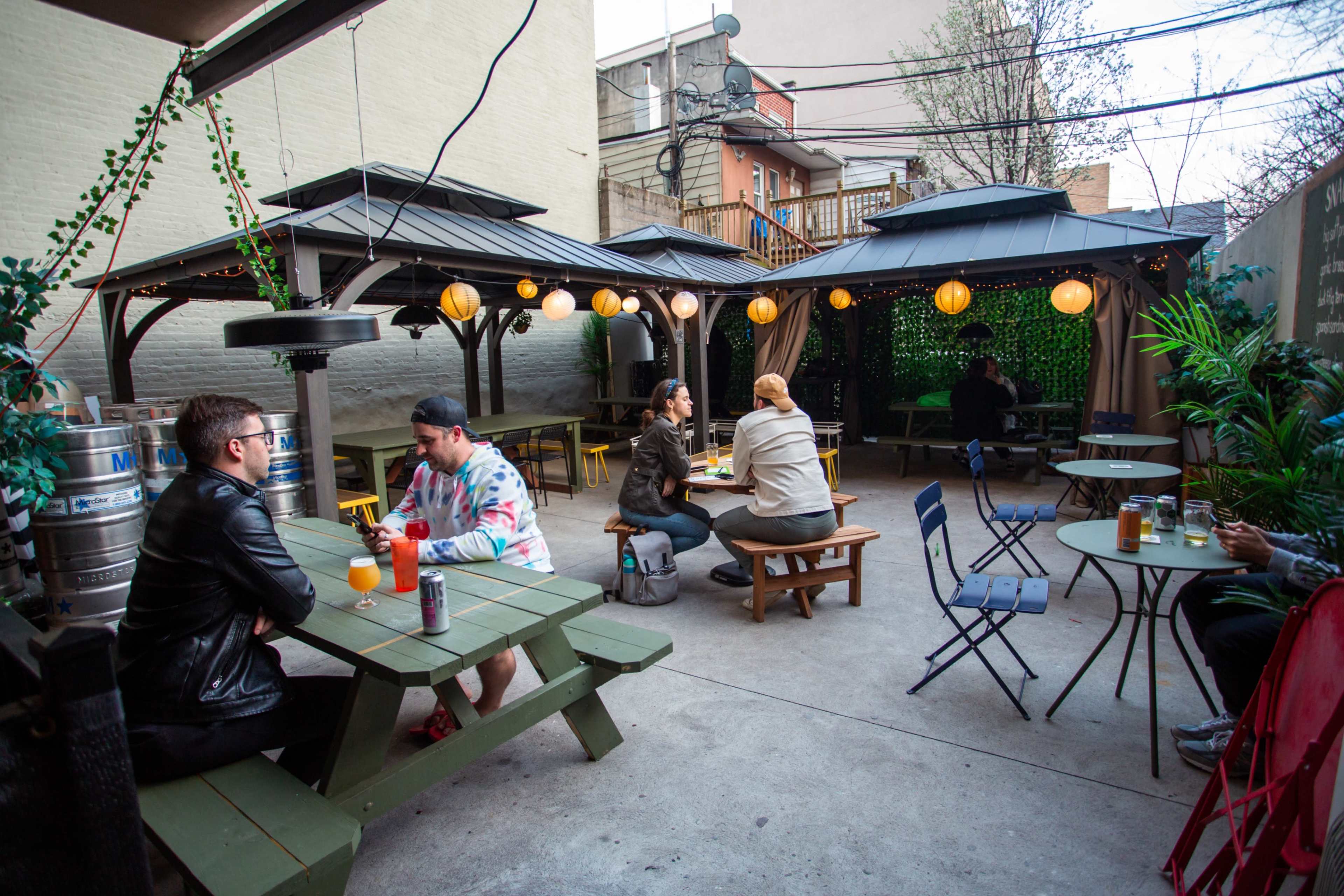 An outdoor patio area with several wooden picnic tables, string lights, and patrons sitting under covered gazebos.