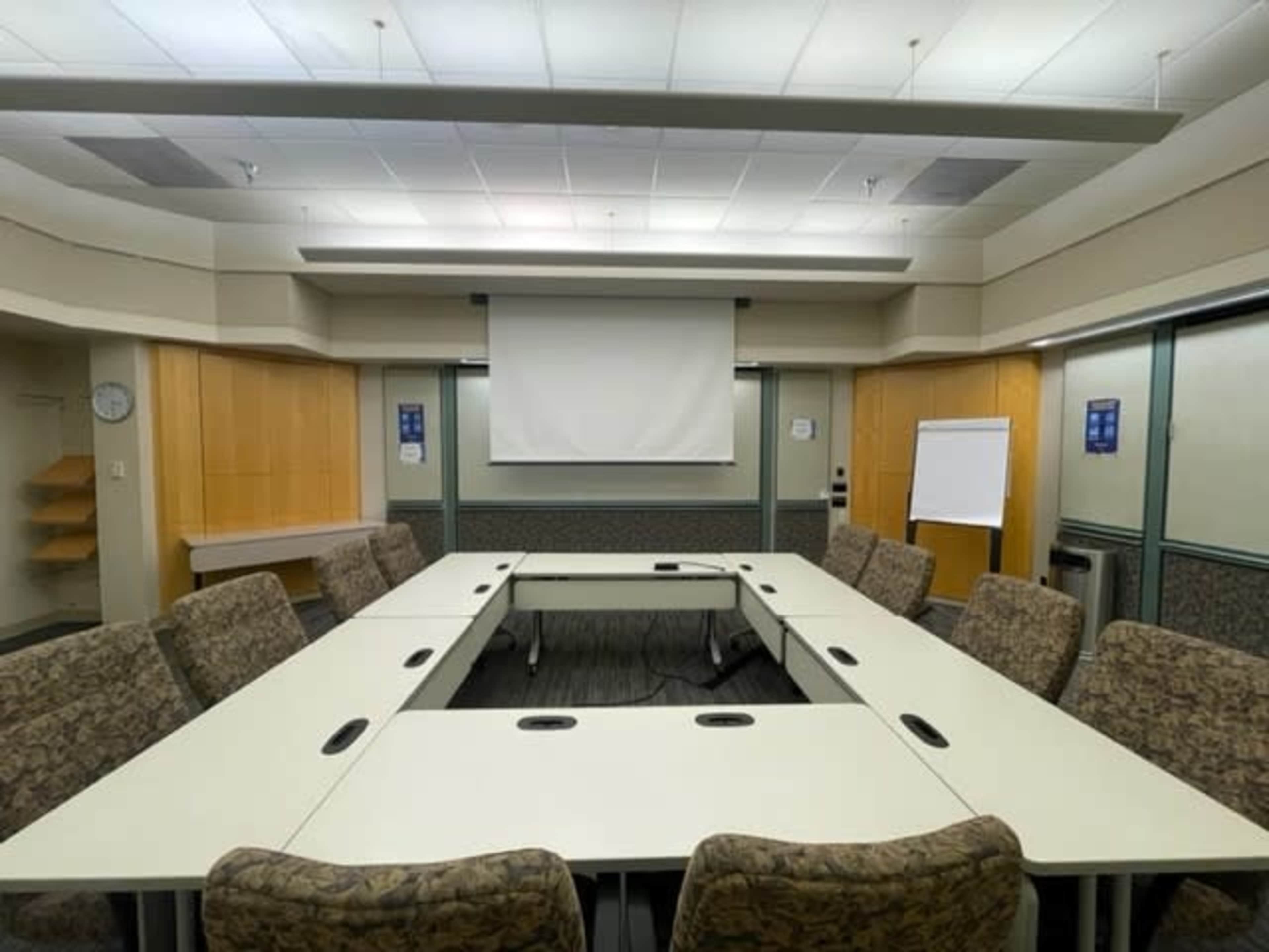 A meeting room with a large rectangular table surrounded by chairs, featuring a projection screen at the front and a whiteboard on the side.