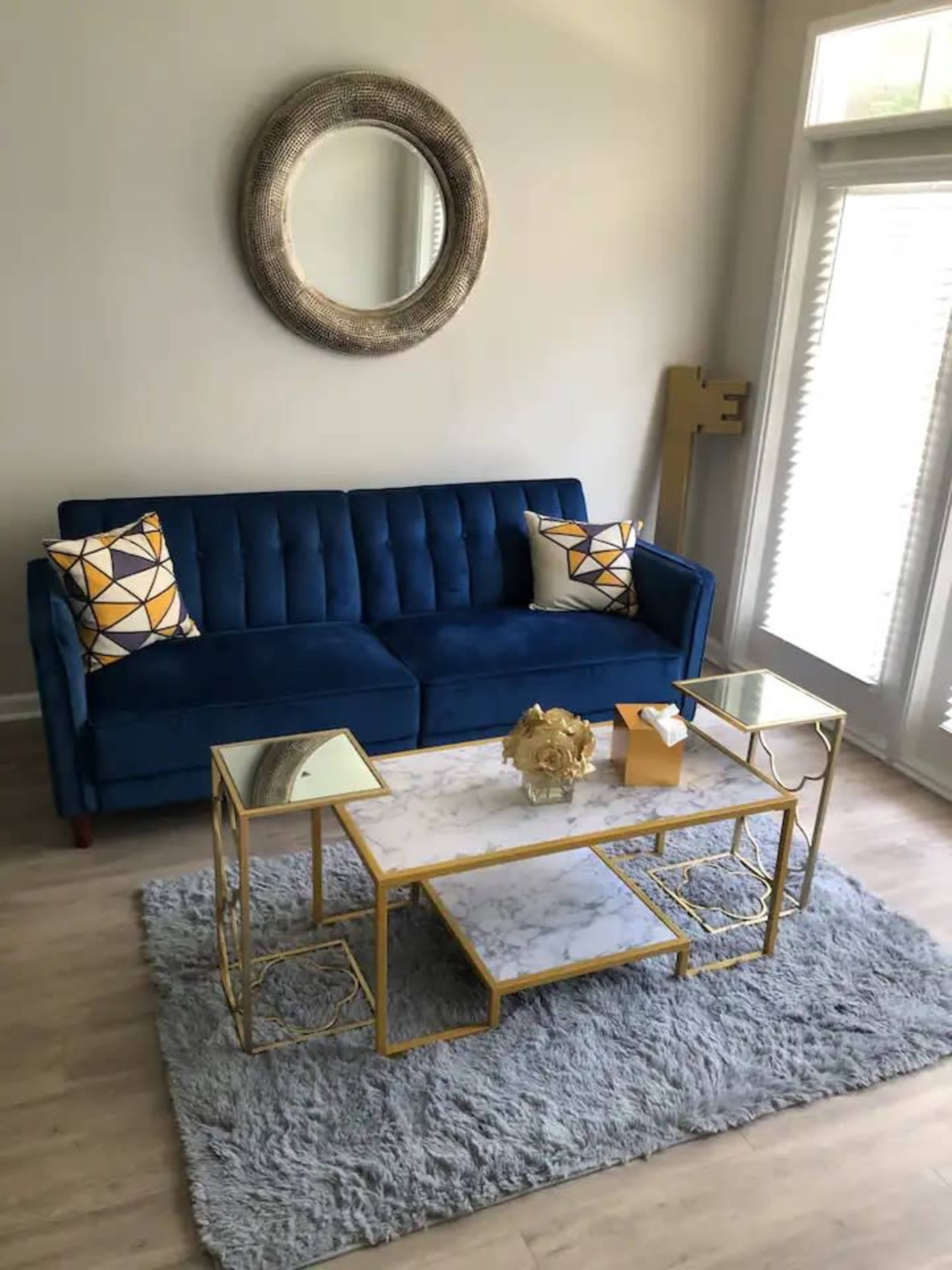 The image shows a modern living room with a blue velvet sofa, decorative pillows, a marble-top coffee table with gold accents, a light gray rug, and large windows allowing natural light to enter.