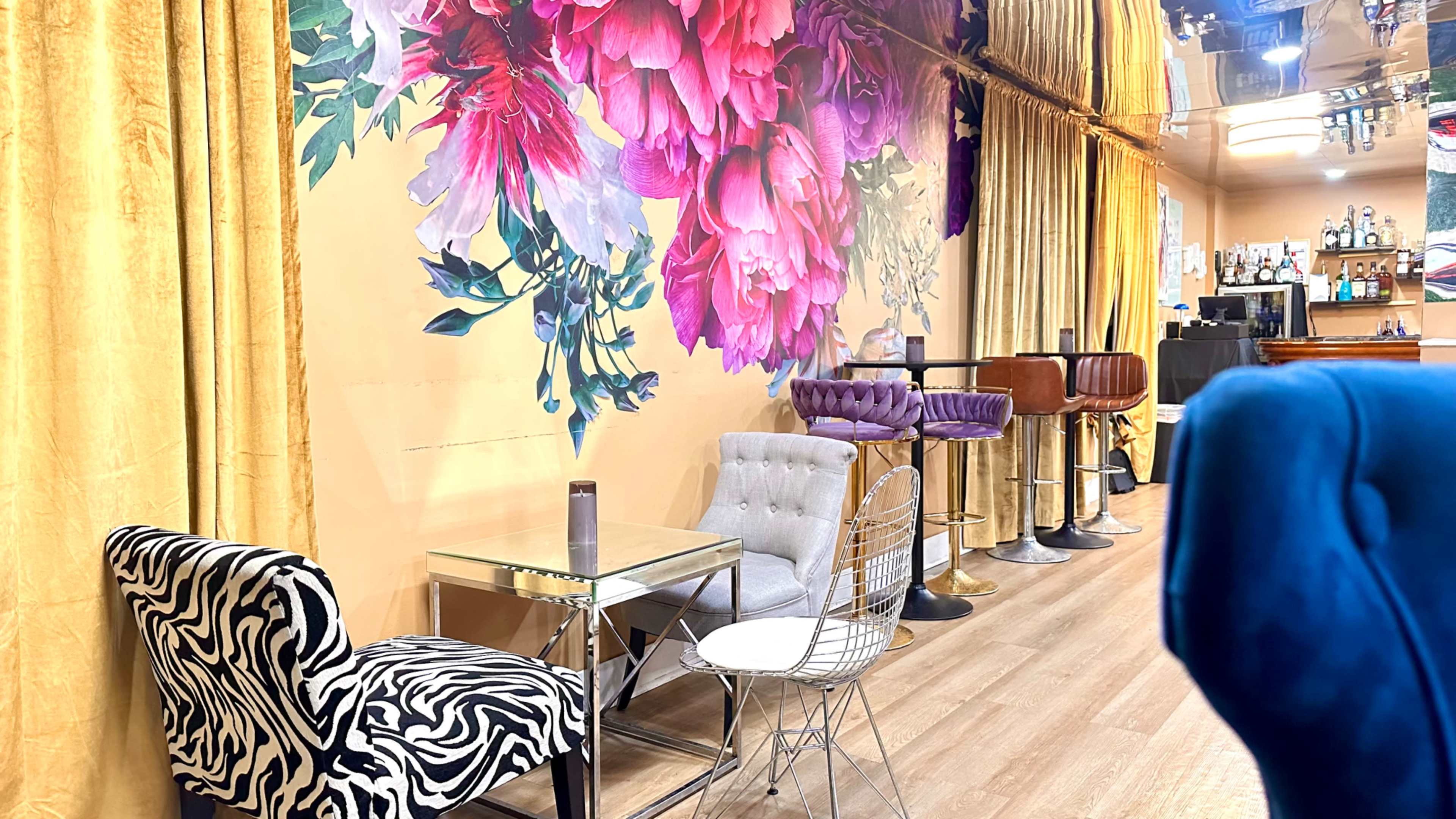 The image shows a vibrant interior of a café or lounge featuring a floral mural on a yellow wall, along with a mix of seating options including a zebra-striped chair, a tufted gray chair, and bar stools.