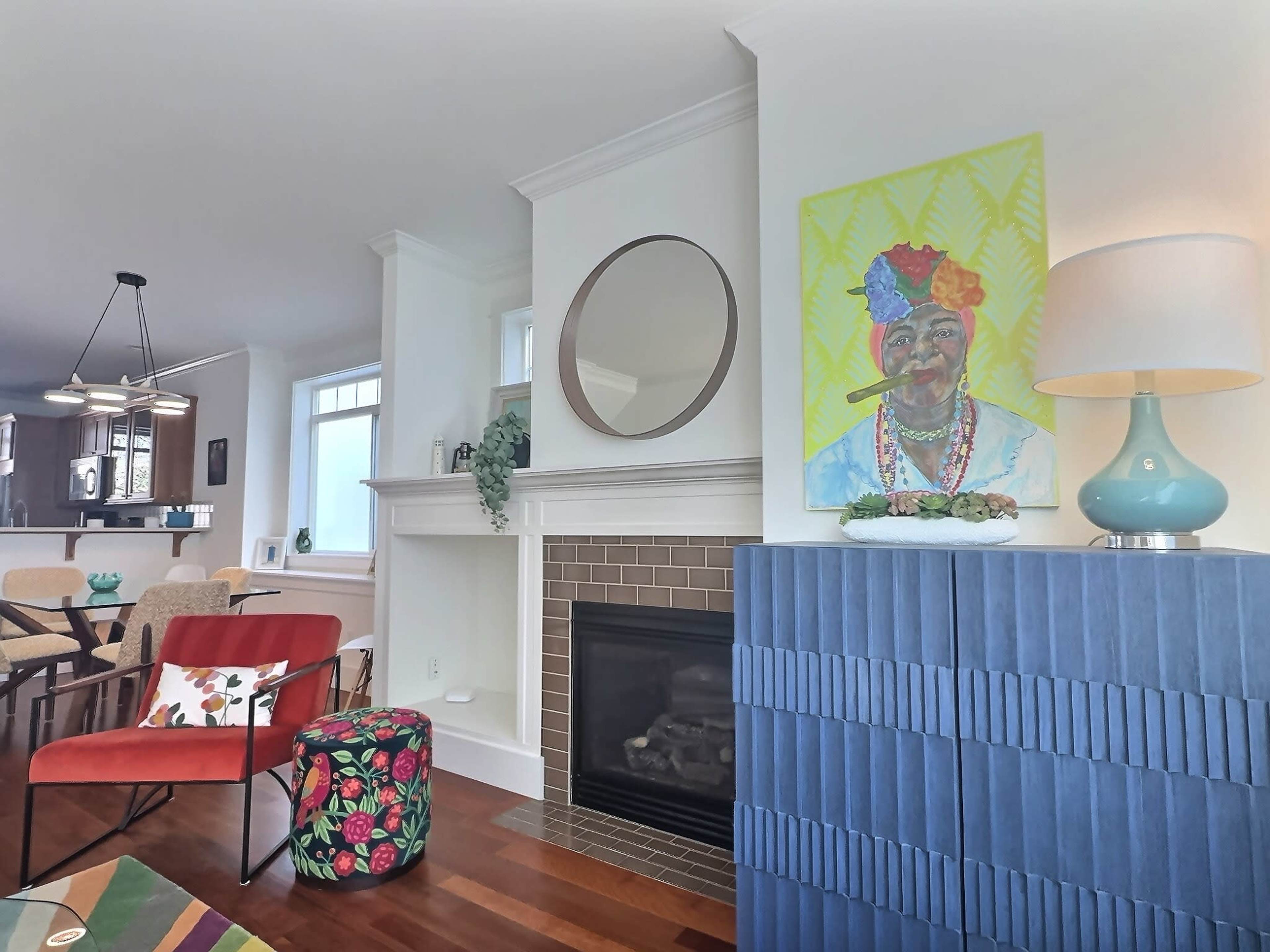 The living room features a modern design with a fireplace, a colorful painting on the wall, and stylish furniture including a red chair and a patterned pouf.