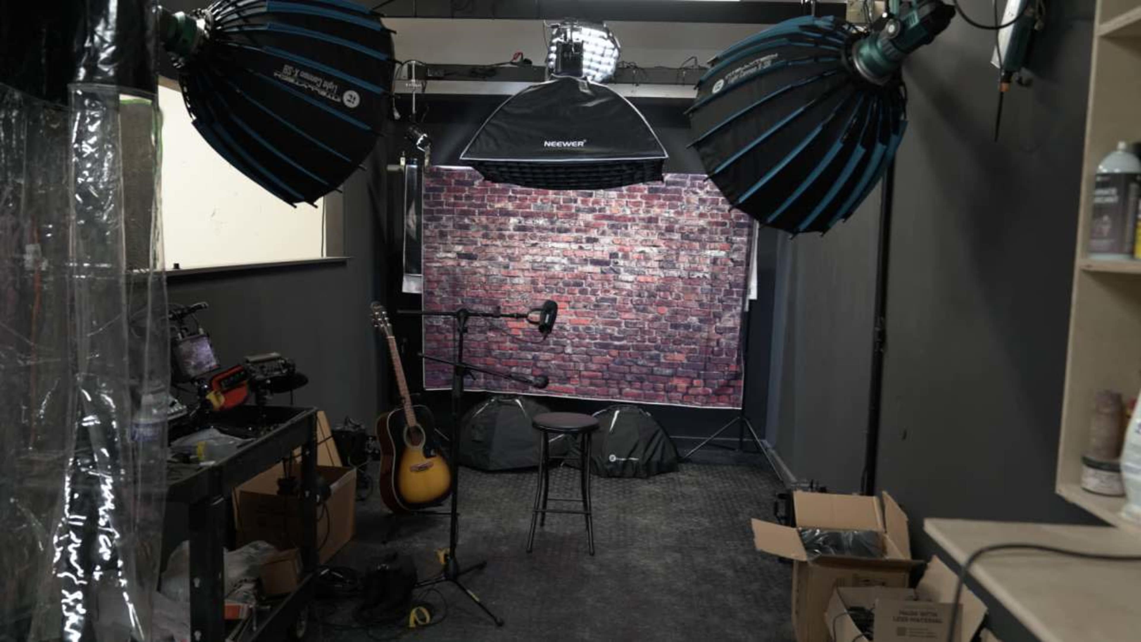 The image shows a dimly lit photo studio featuring a brick backdrop, a stool, an acoustic guitar, and multiple lighting setups.