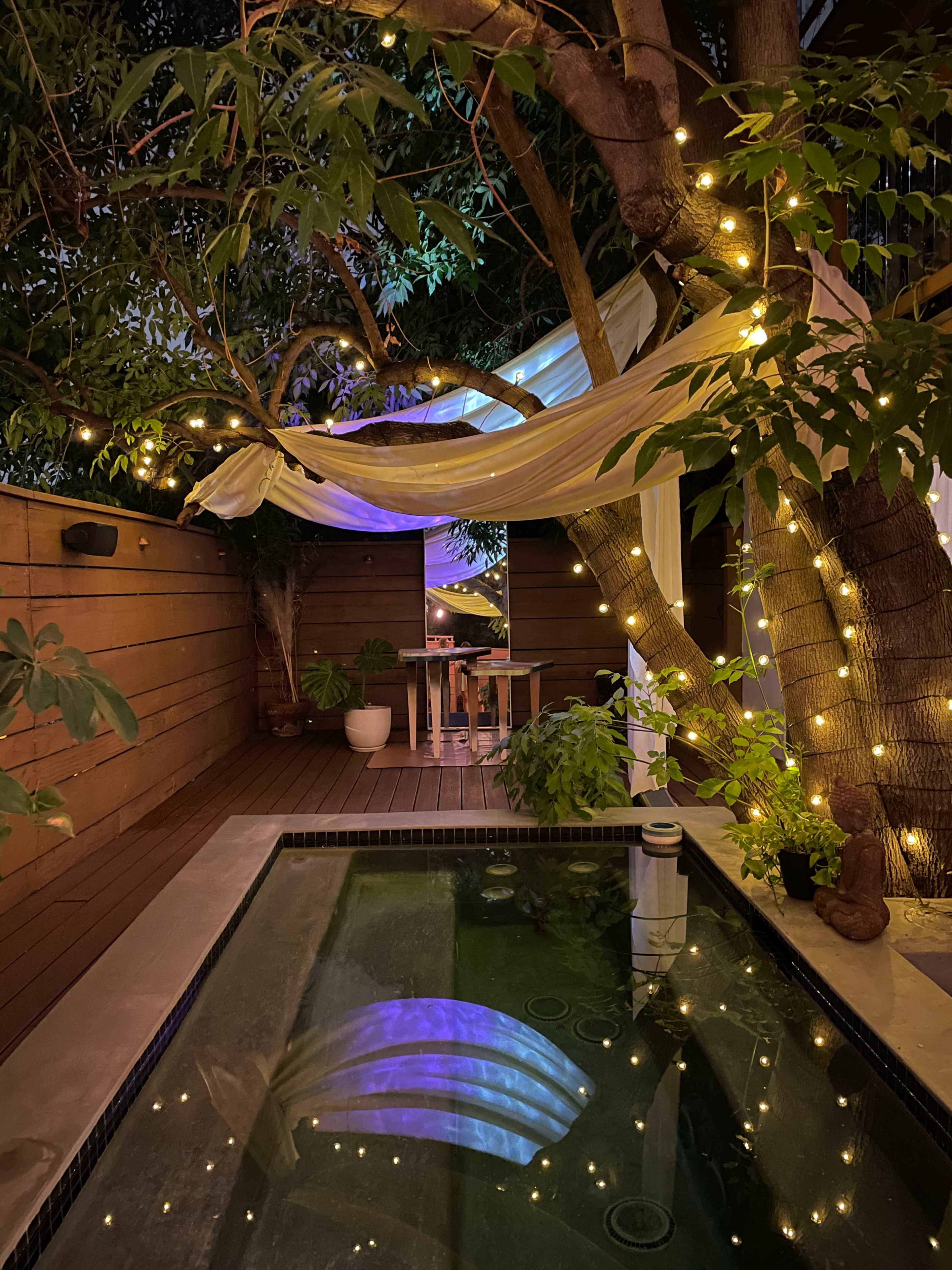 A small, illuminated outdoor space features a pool surrounded by greenery, with a dining table beneath draped fabric and string lights in the trees.