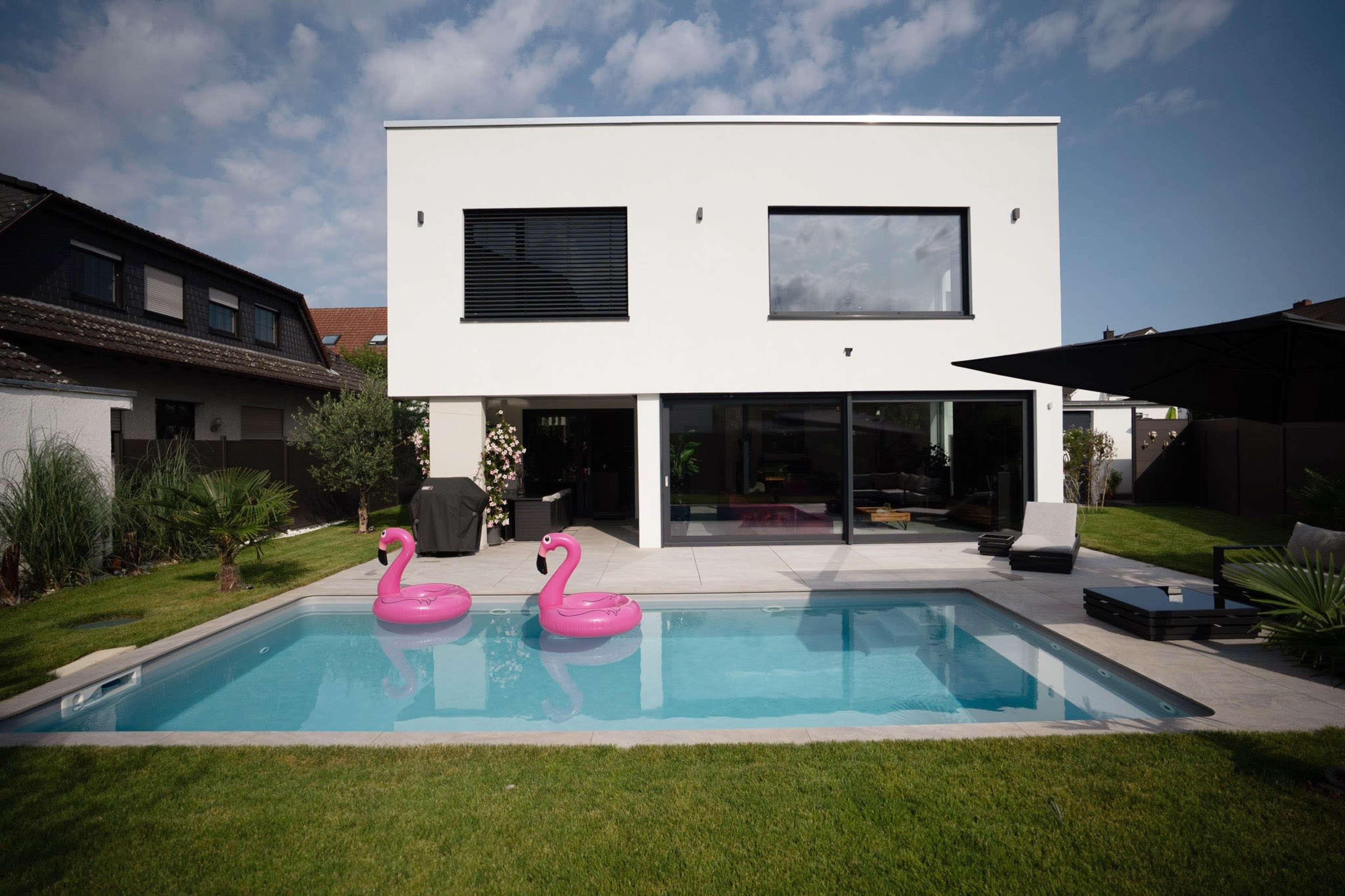 The image shows a modern two-story house with large windows overlooking a swimming pool that features two pink inflatable flamingos.