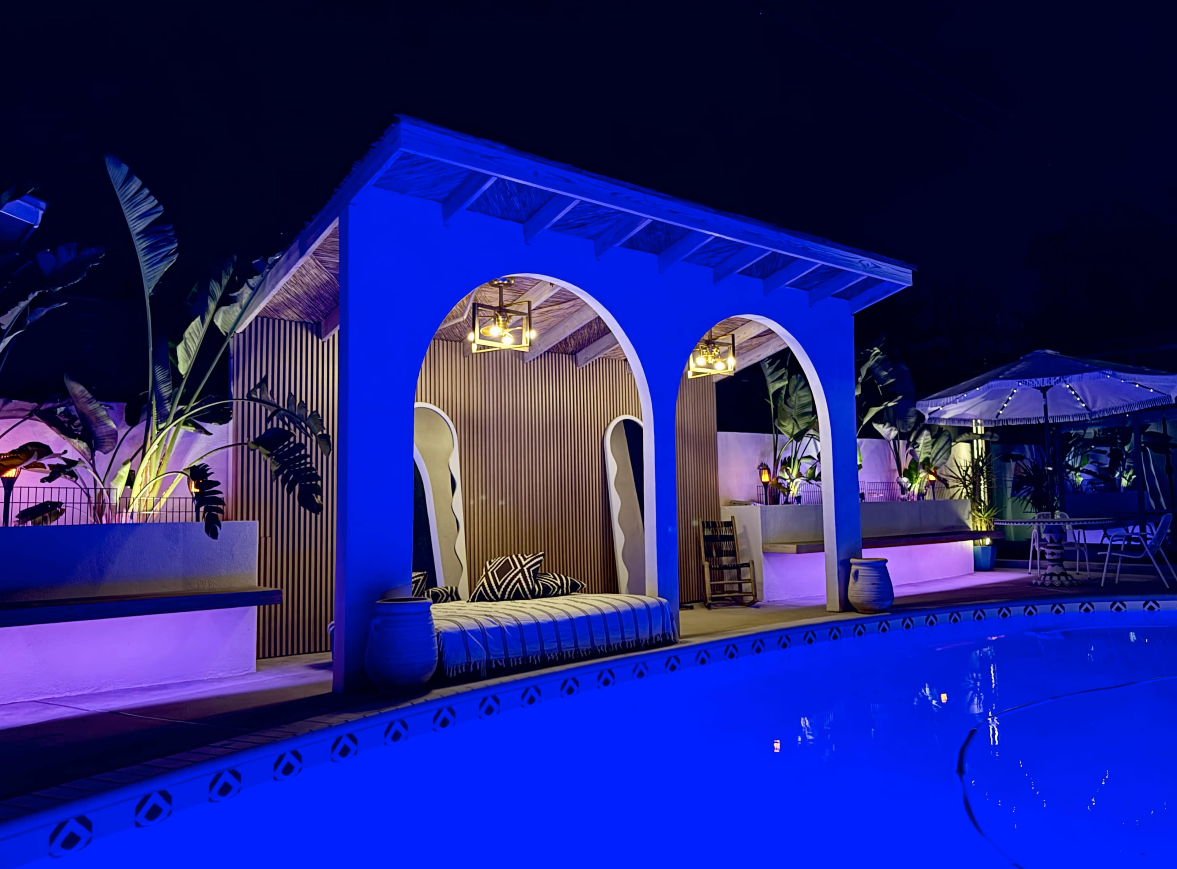 A brightly lit, modern outdoor space features a pool surrounded by tropical plants, decorative lighting, and lounge areas with arches.