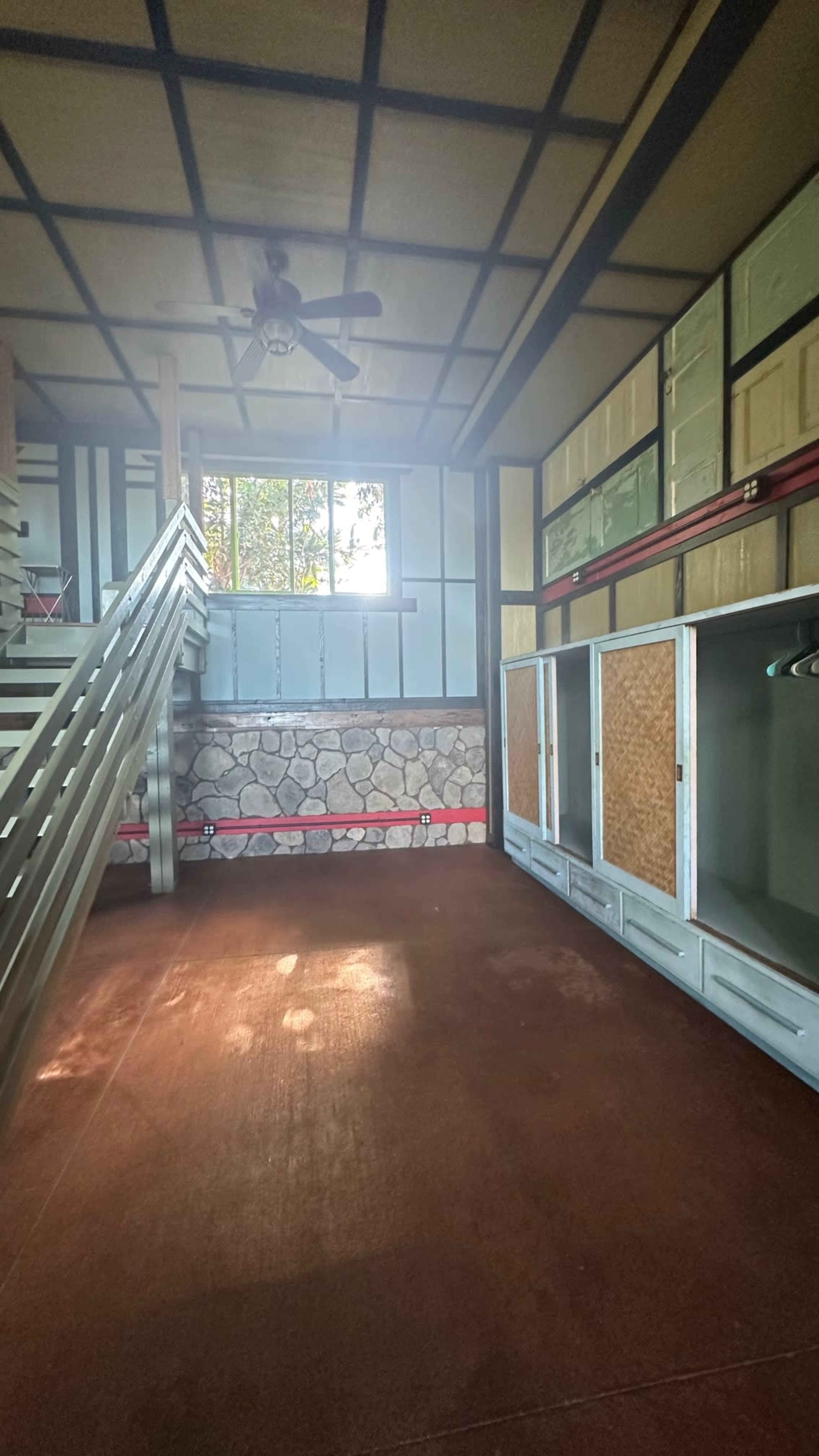 A sparse interior space with wooden stairs on the left, a stone wall on the bottom, and large windows allowing sunlight to enter.