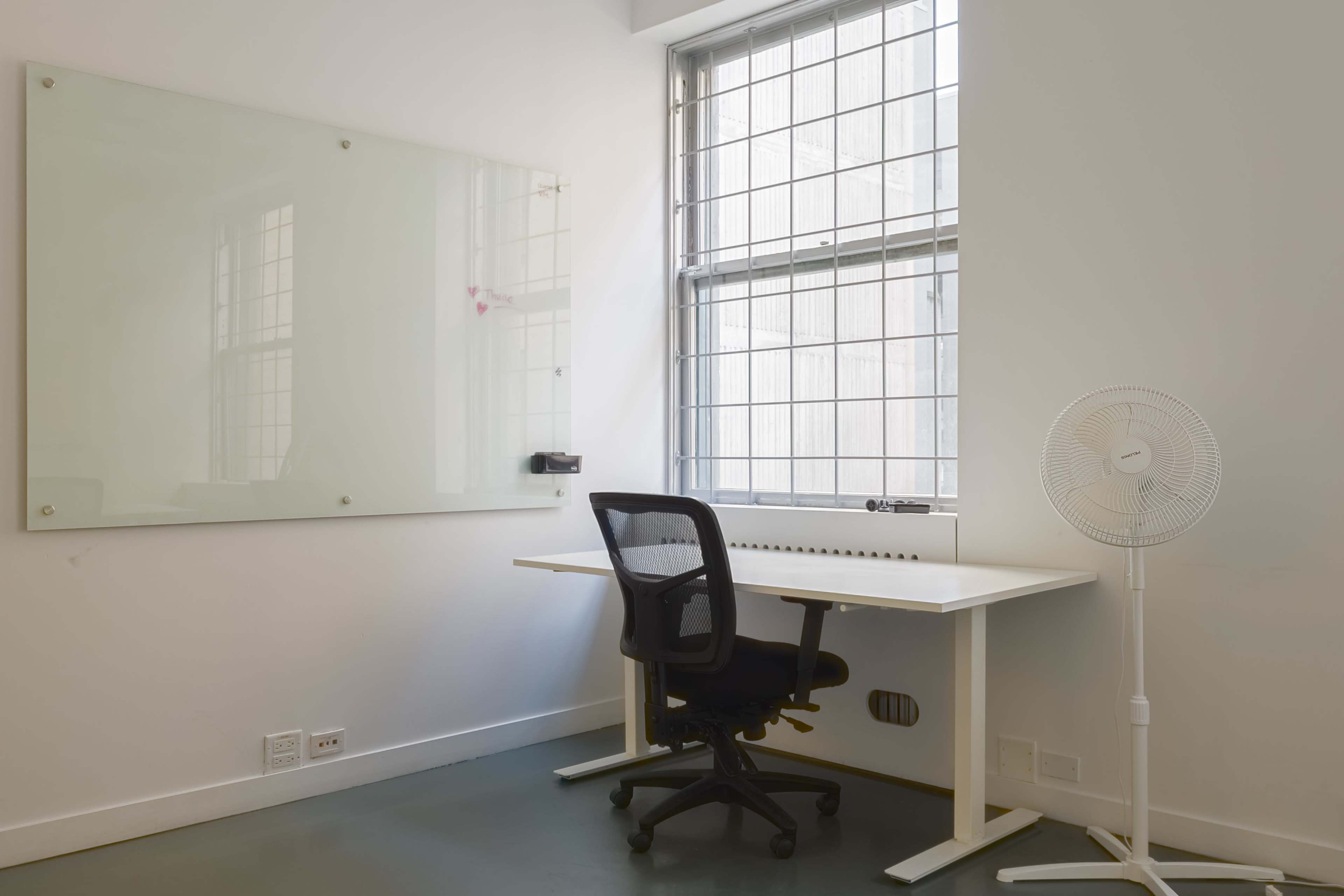The image shows a minimalistic office space with a white desk, a black office chair, a standing fan, and a large window with light pouring in.