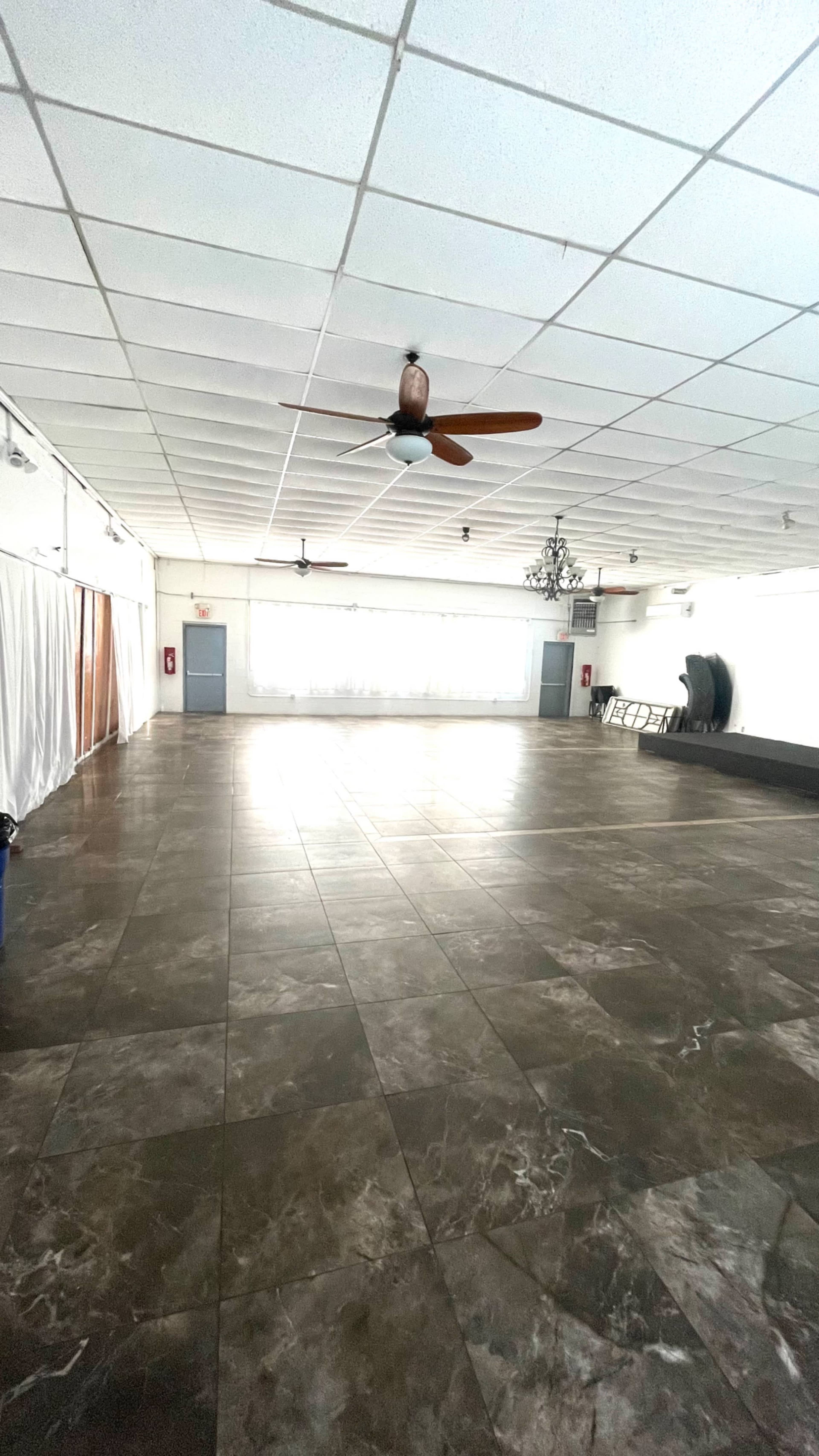 The image shows a large, empty room with tiled flooring, ceiling fans, and a few furnishings along the walls.