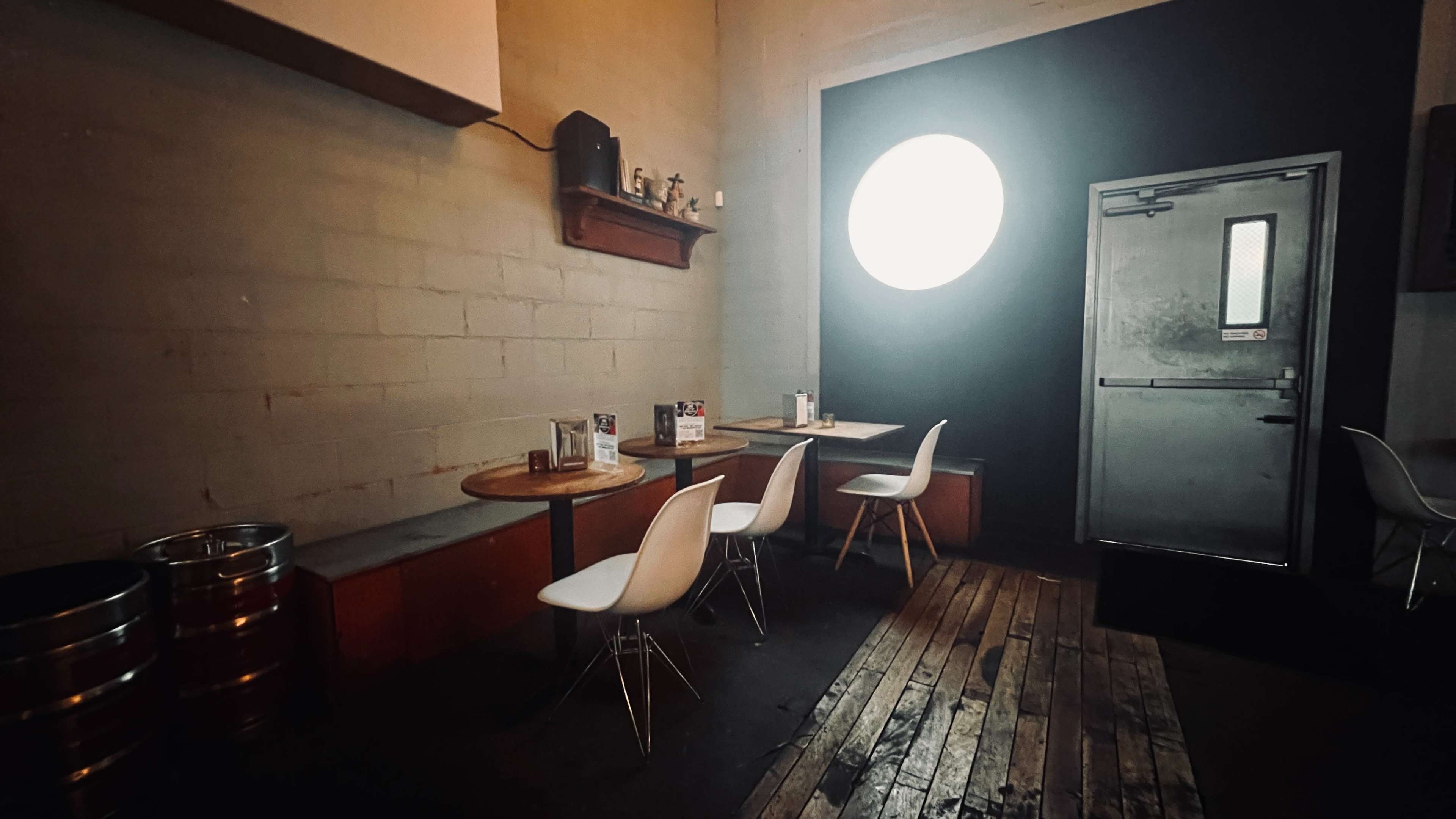 A dimly lit café interior with wooden flooring, a large round window on the black wall, and several white chairs around small tables.