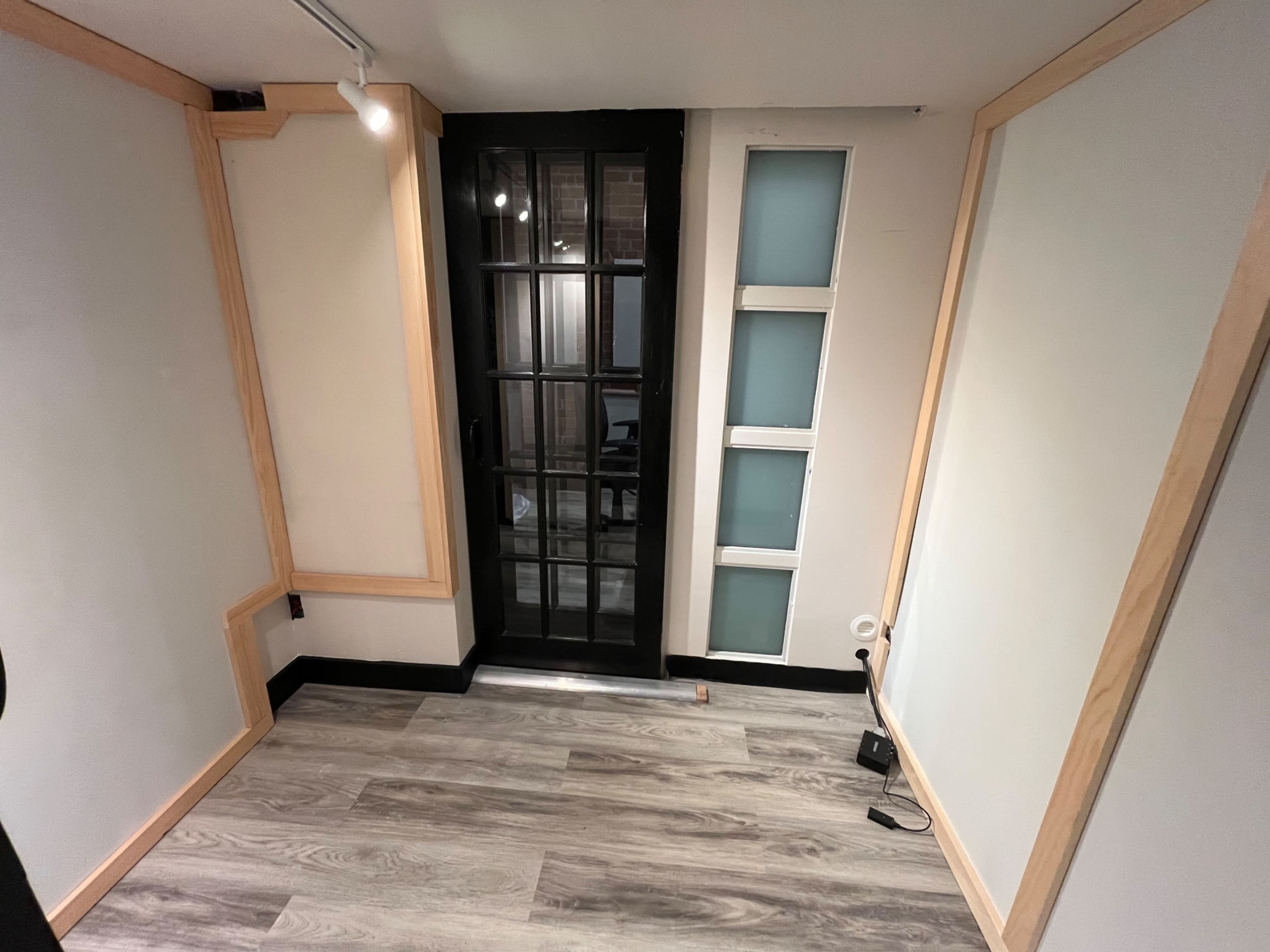 The image shows a small room with hardwood flooring, featuring a glass door leading outside and sections of wall with a light wood trim.