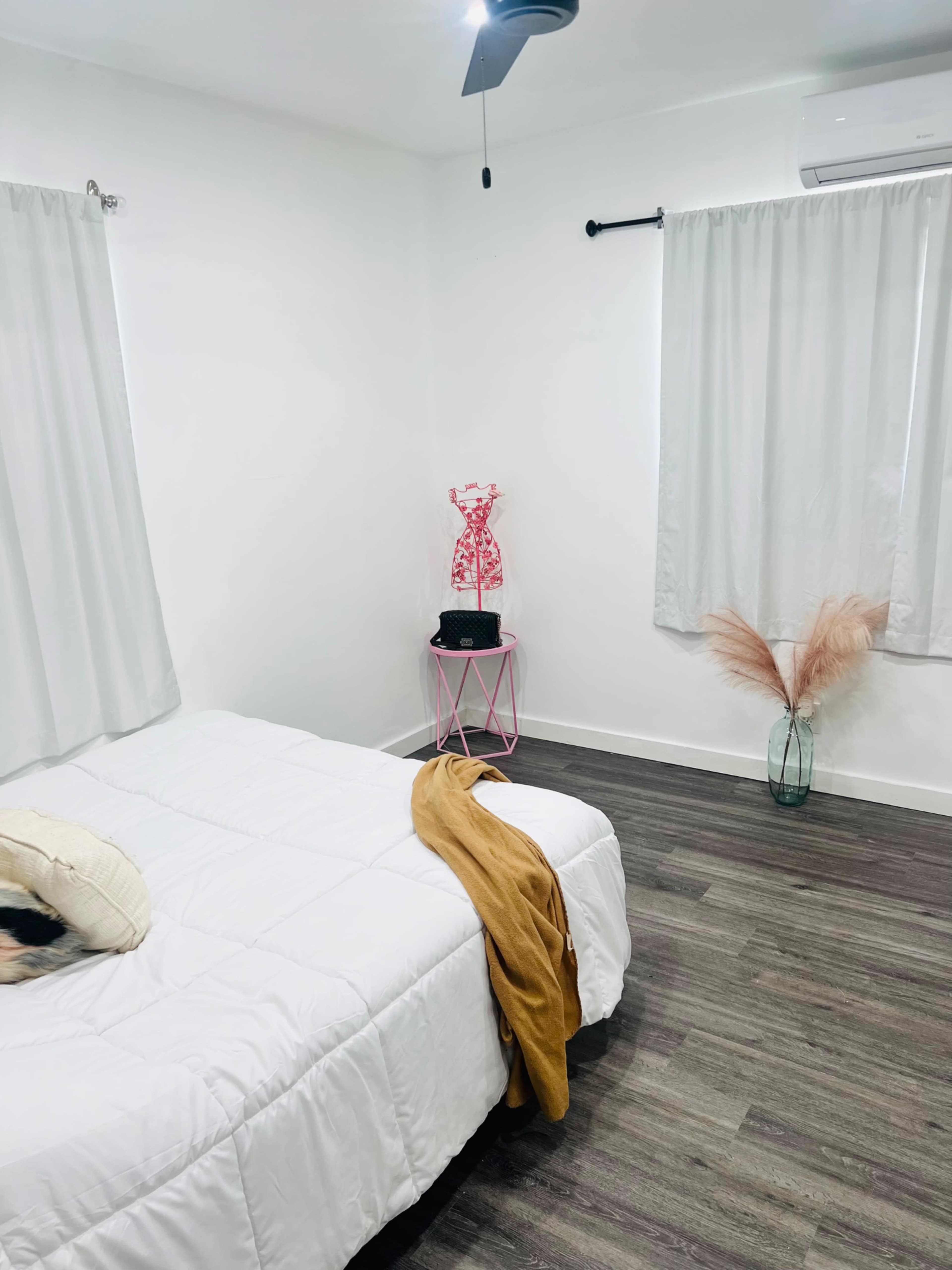 A cozy bedroom features a white bed, a pink side table with a decorative figure, and two sheer curtains framing a window.