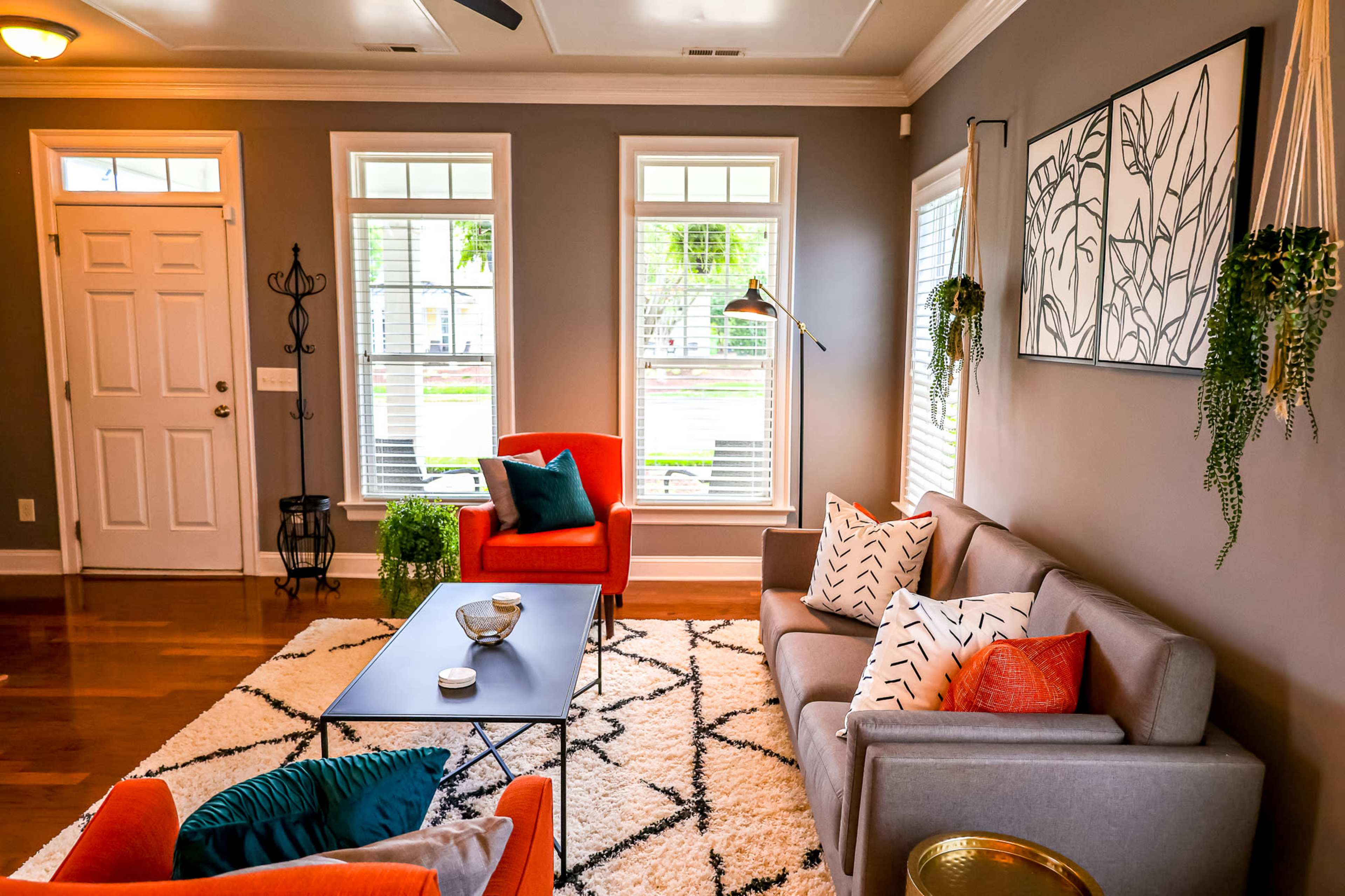 The living room features a gray wall color, a mix of orange and gray furniture, large windows, and a patterned rug.