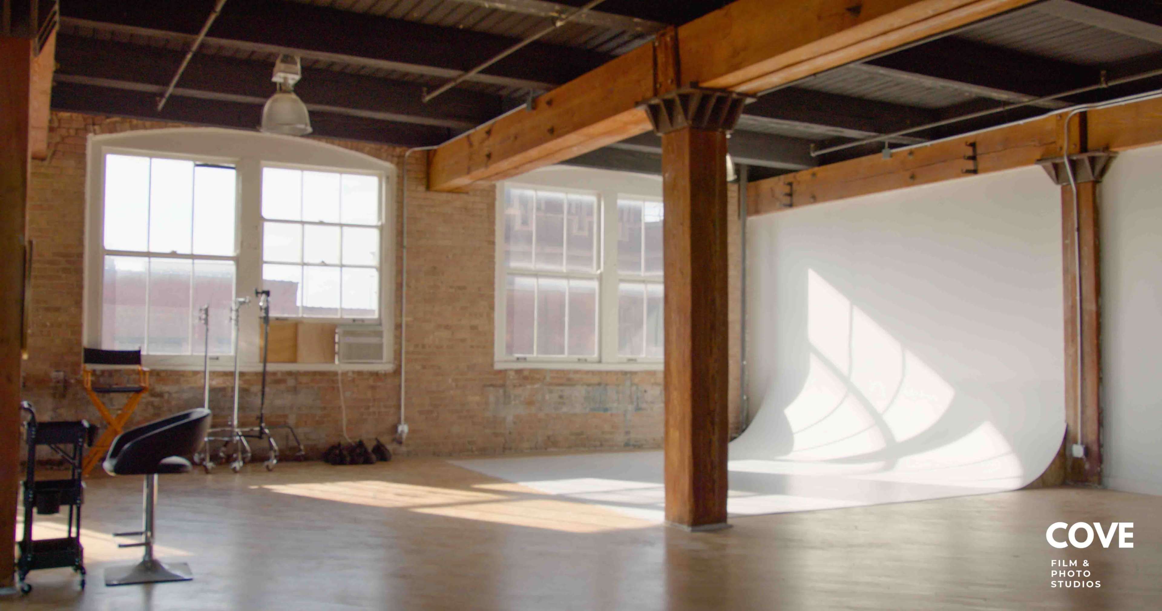The image shows an empty studio space with large windows, wooden beams, a white backdrop, and film equipment in the corner.