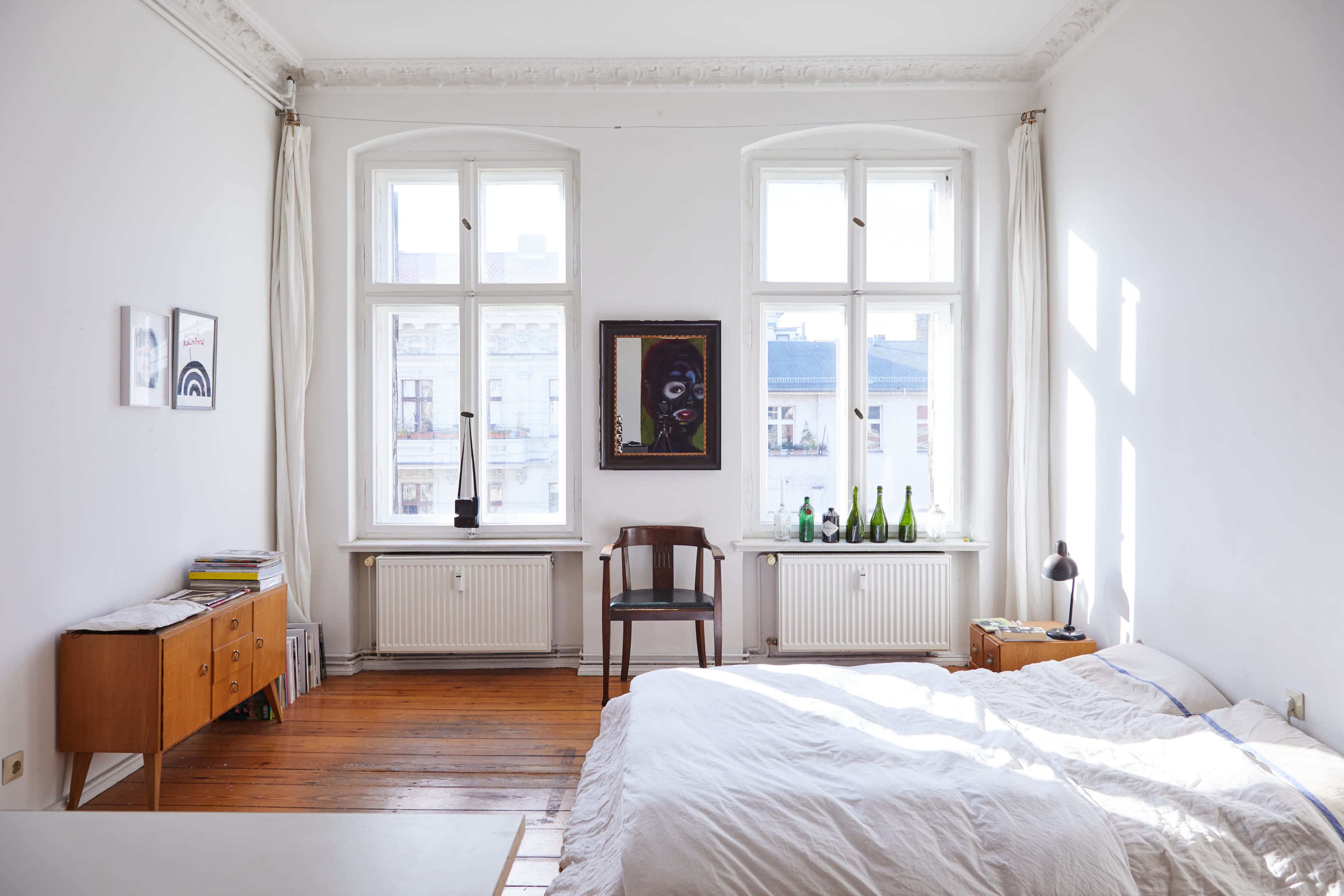 The bedroom features a bed with white bedding, a wooden chair, a simple dresser, and large windows letting in natural light.