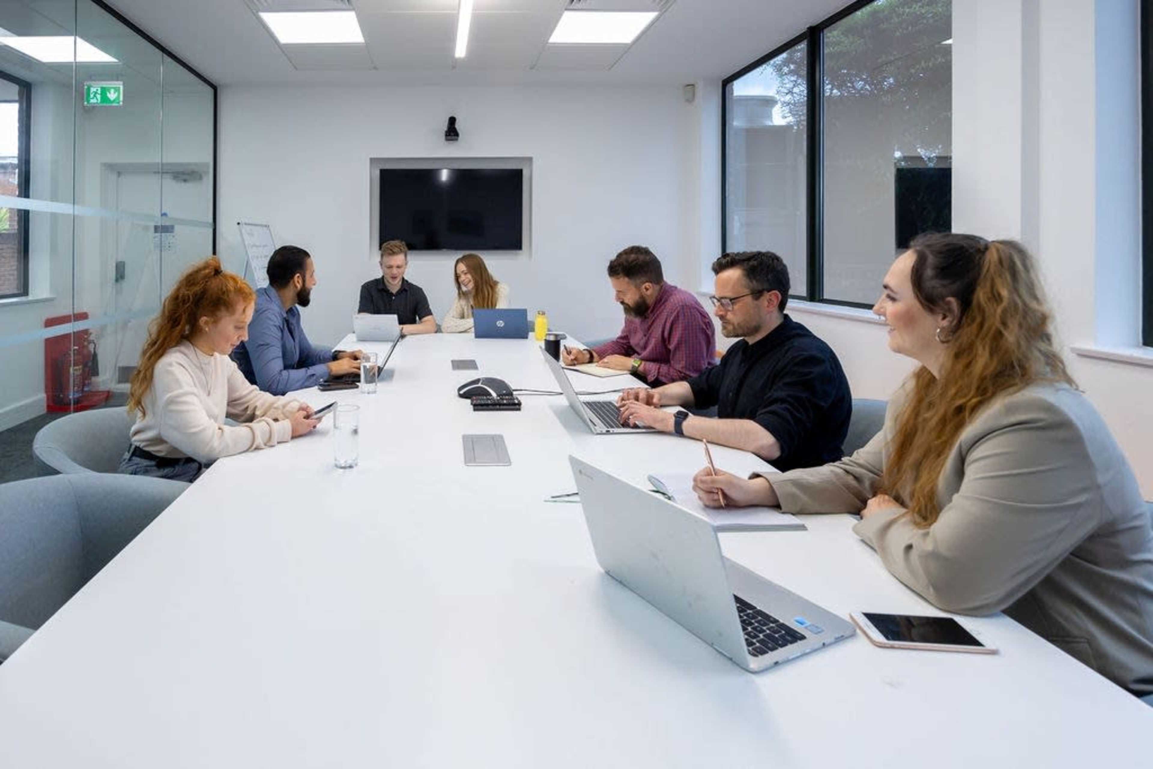 A group of six people is seated around a long white table in a conference room, engaged in discussions and working on laptops.