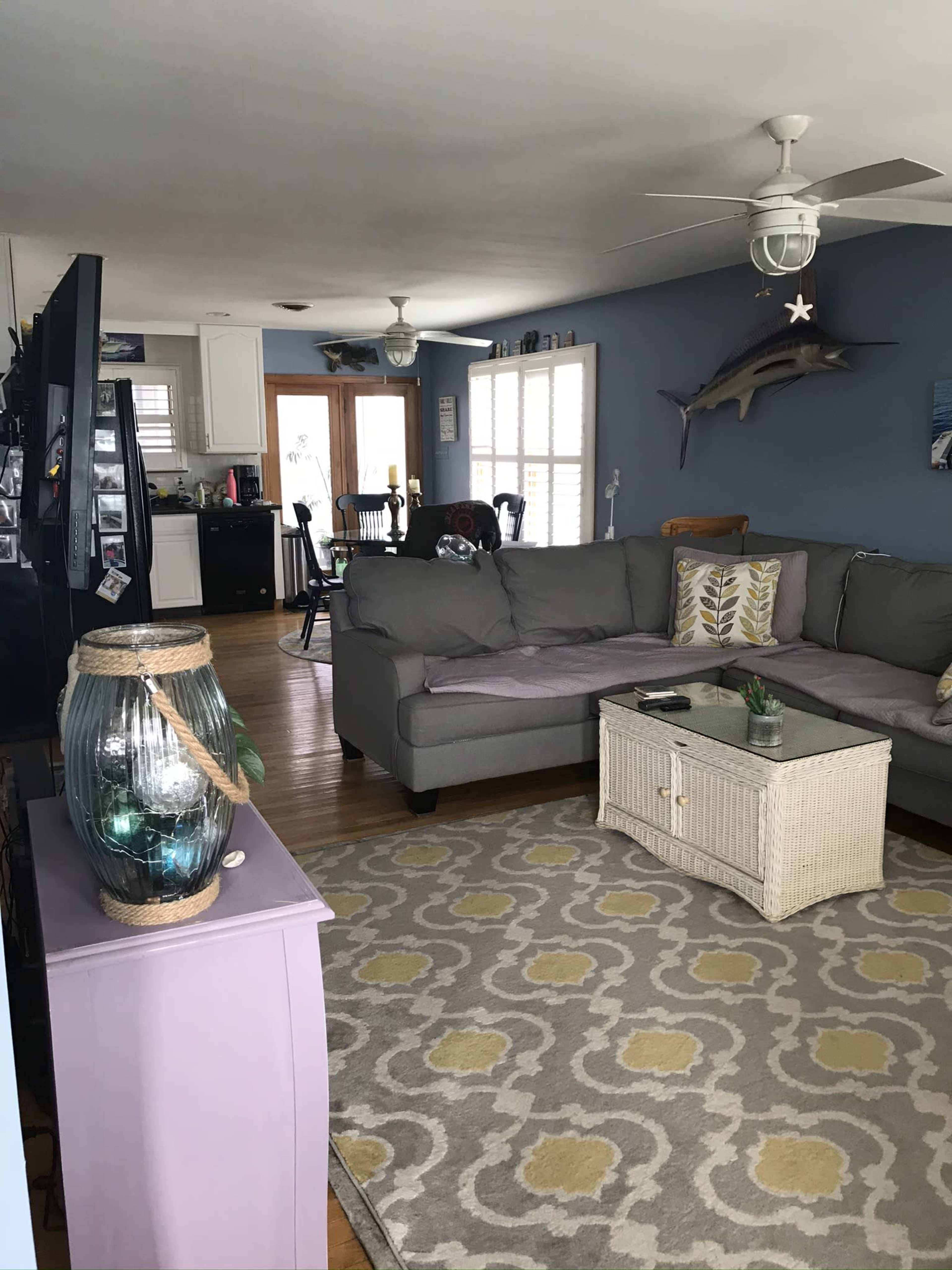 A living room with a gray sectional sofa, a white woven coffee table, and a light-colored area rug, leading to a kitchen area in the background.