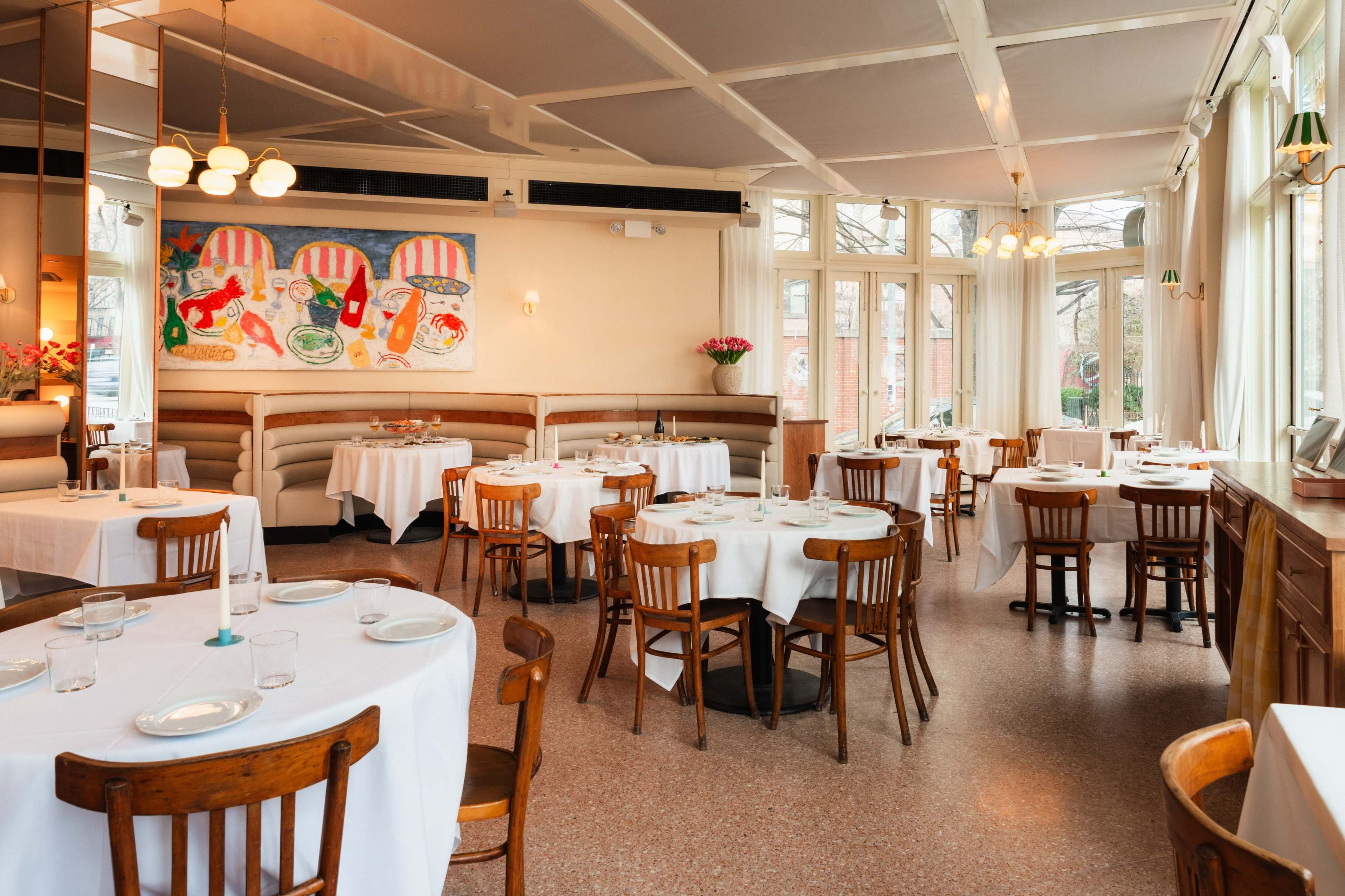 The image shows a restaurant interior with round tables covered in white tablecloths, wooden chairs, and large windows allowing natural light.