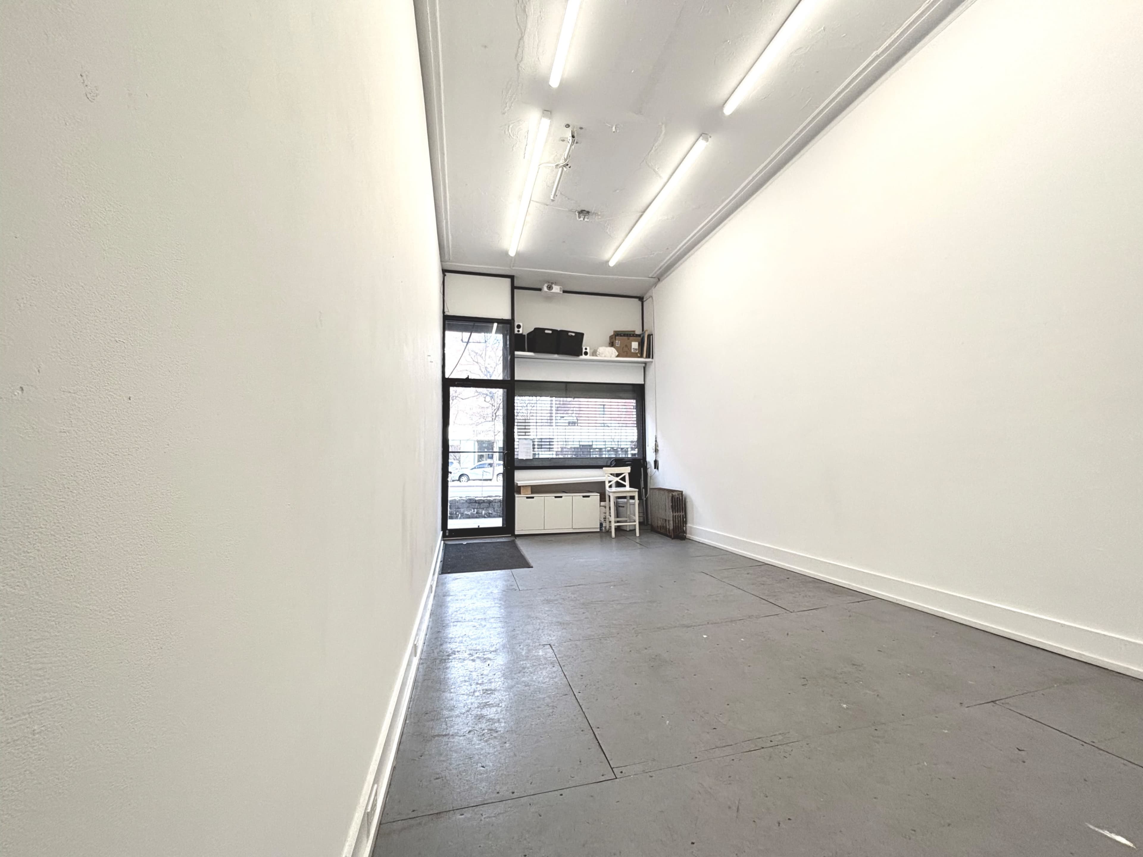 The image shows an empty, well-lit interior space with white walls and a concrete floor, featuring a large window at the front.
