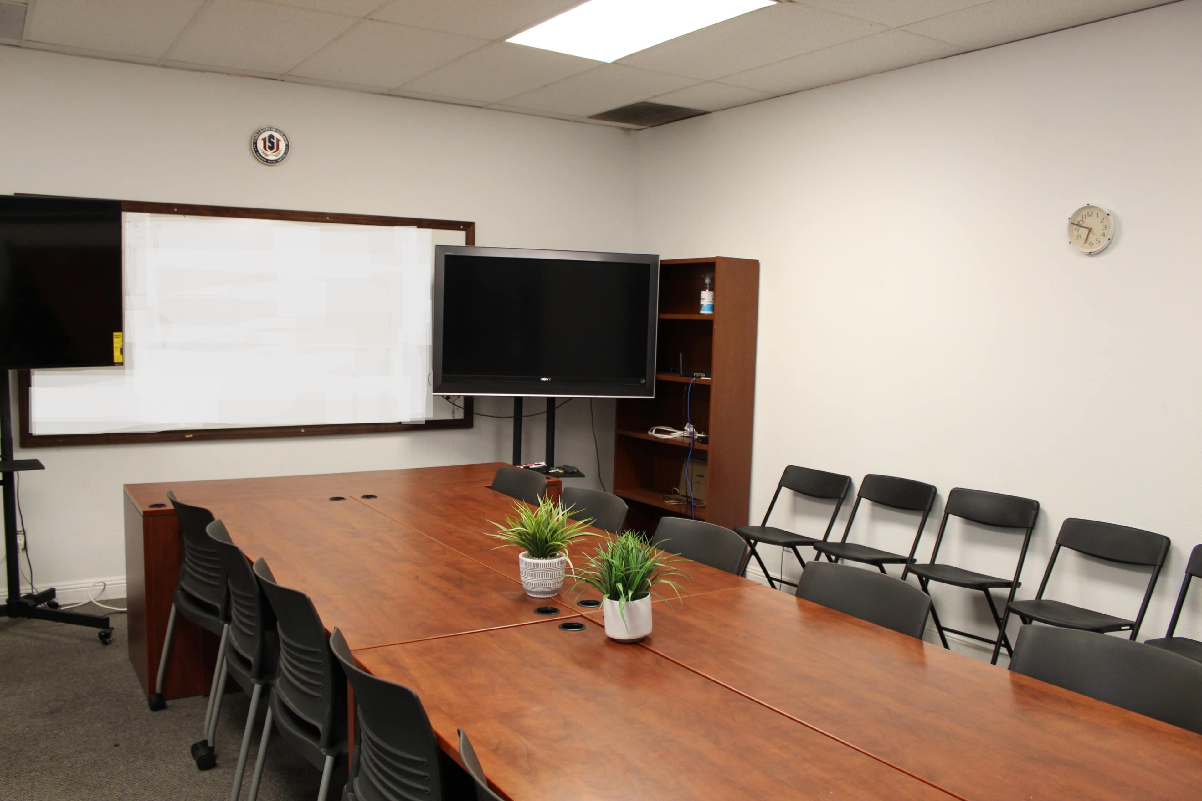 A conference room features a long wooden table surrounded by black chairs, with a large TV and whiteboard on one wall.