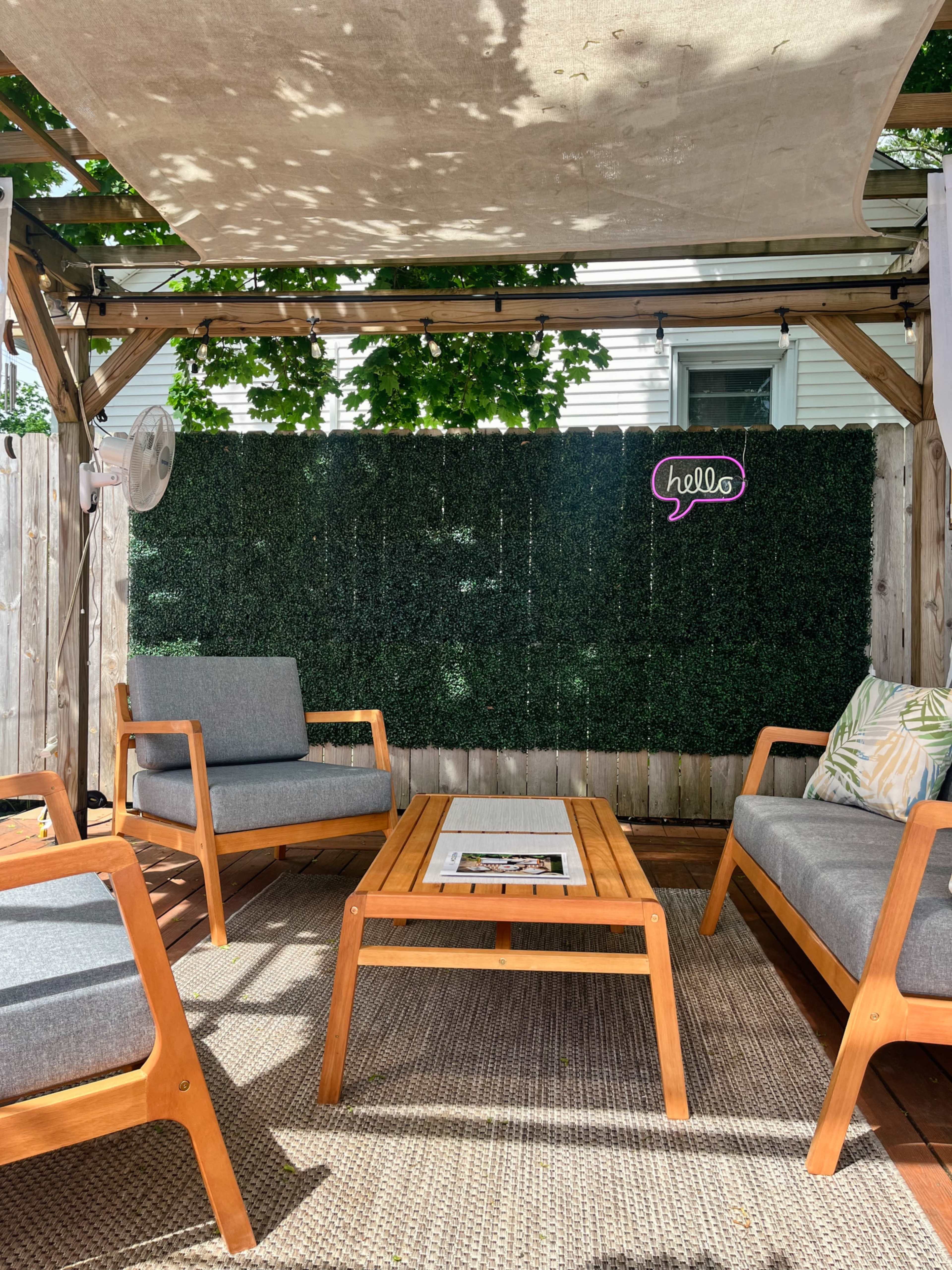 The image shows a shaded outdoor seating area with two wooden chairs and a coffee table, surrounded by a greenery backdrop and a "hello" sign on the wall.