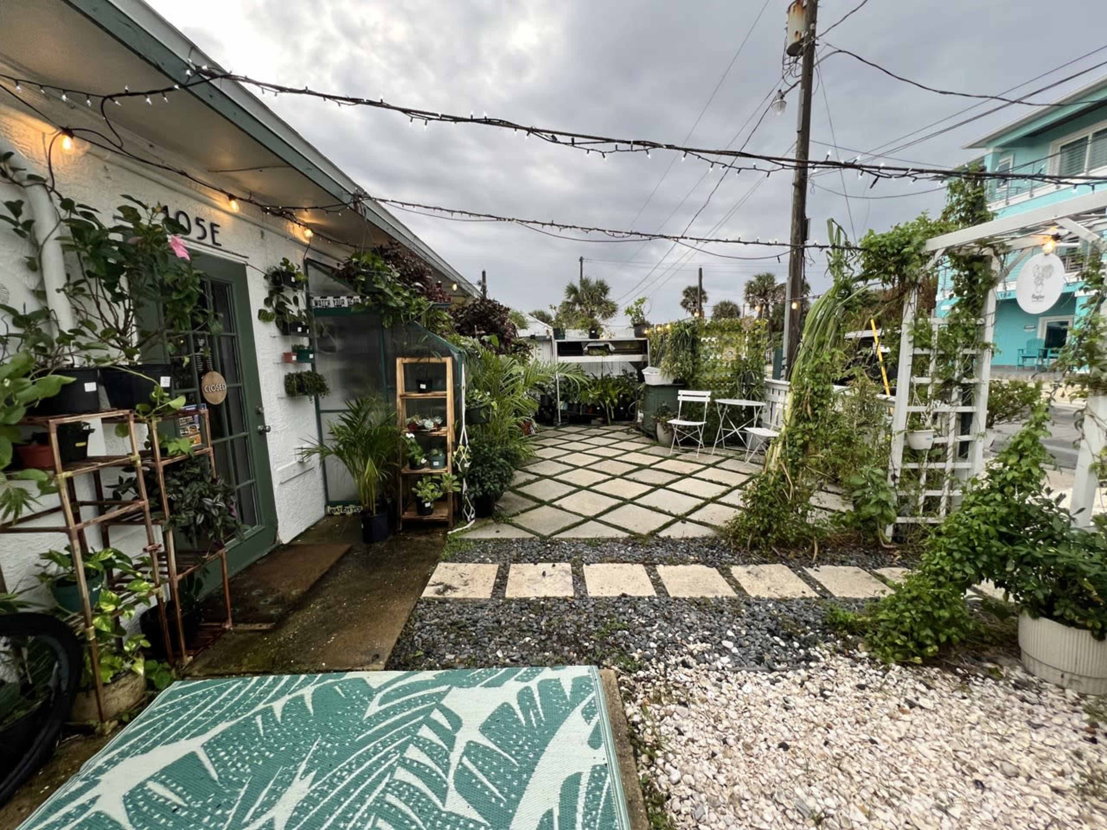 The image shows a garden area with stone pathways, greenery, and a cozy seating arrangement in front of a building under a cloudy sky.