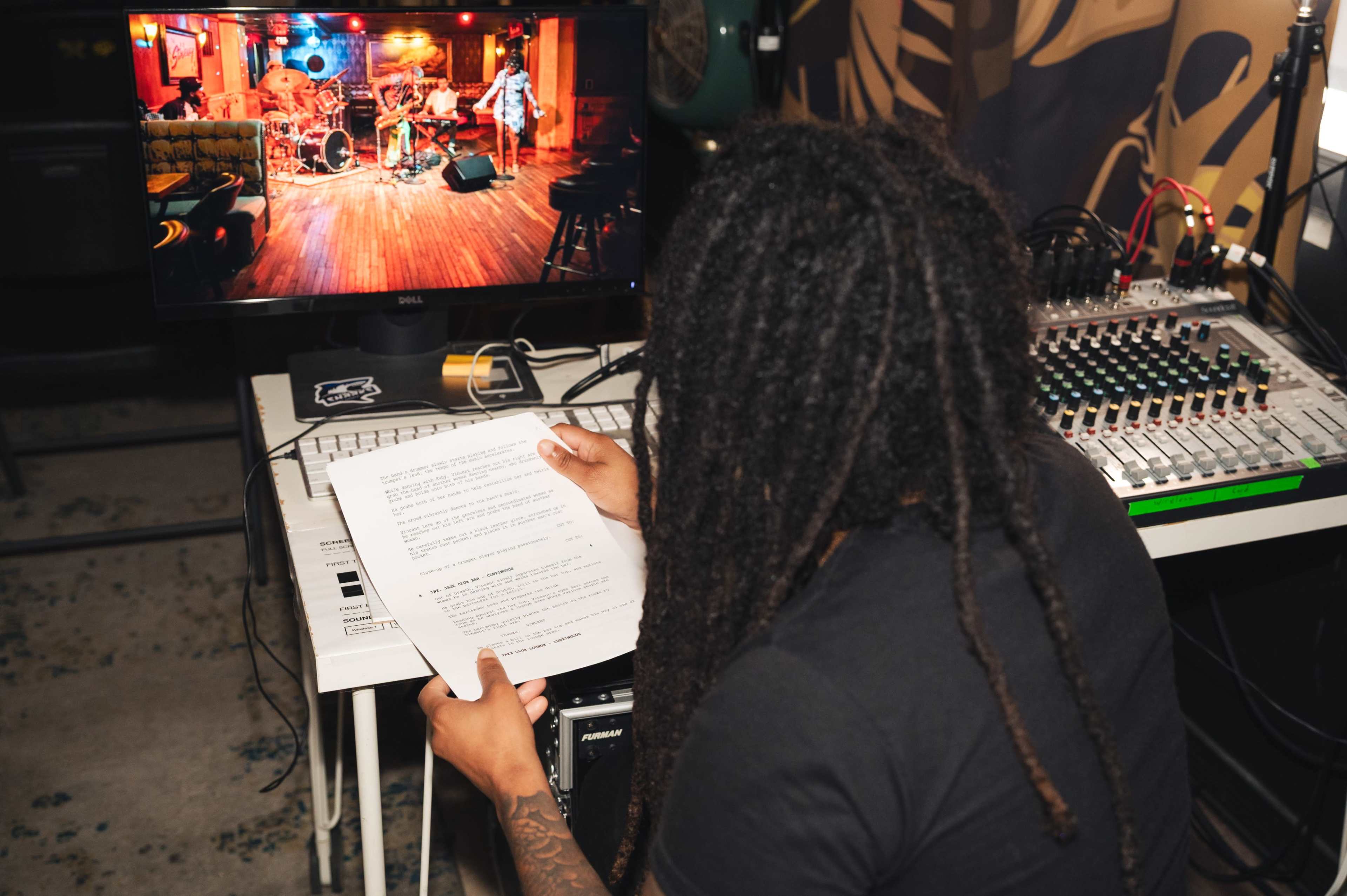 A person with long hair reviews a document while looking at a monitor displaying a live music performance in a vibrant venue.