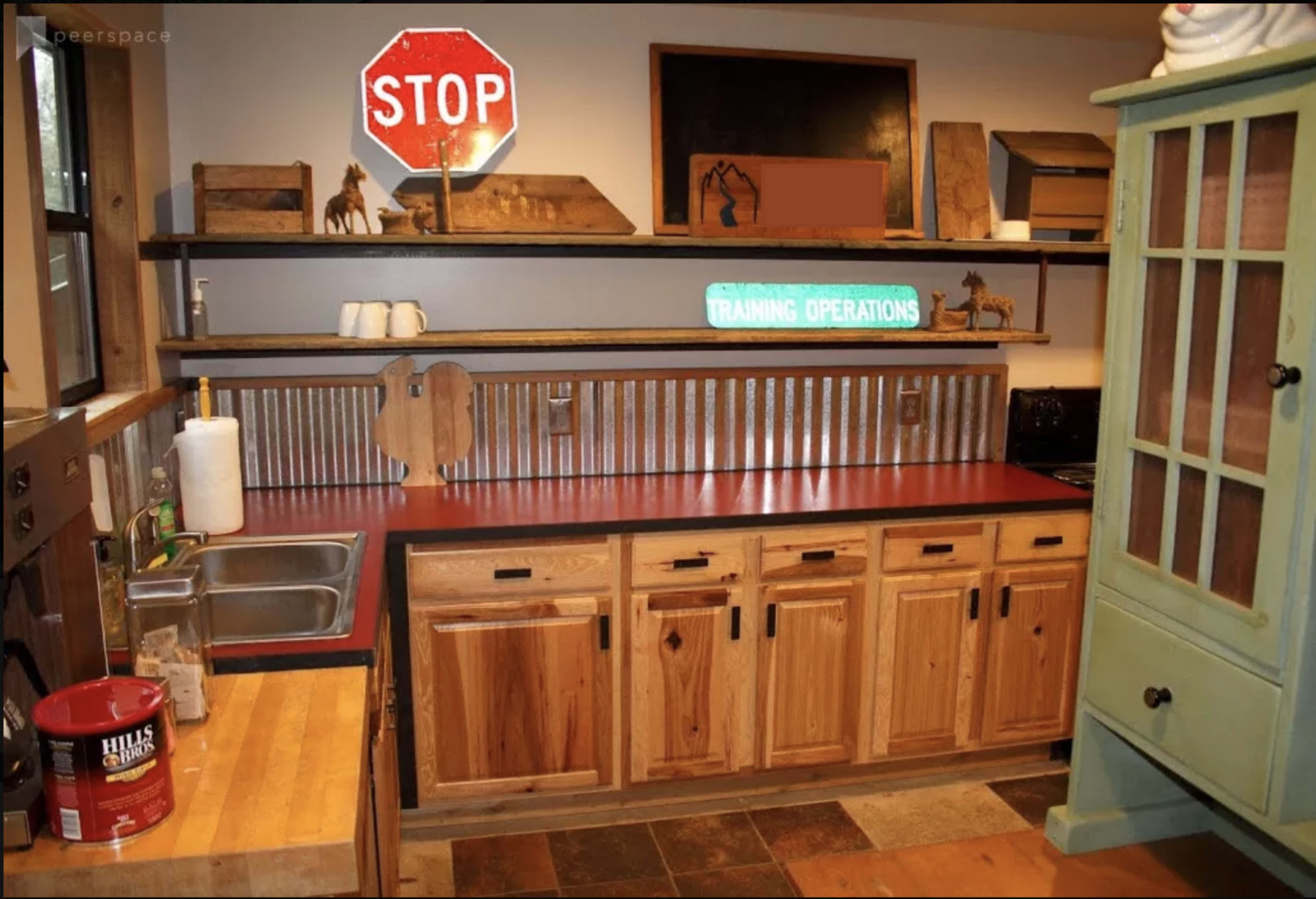 A rustic kitchen featuring wooden cabinets, a stainless steel sink, a red countertop, and a mix of decorative signs and shelves.