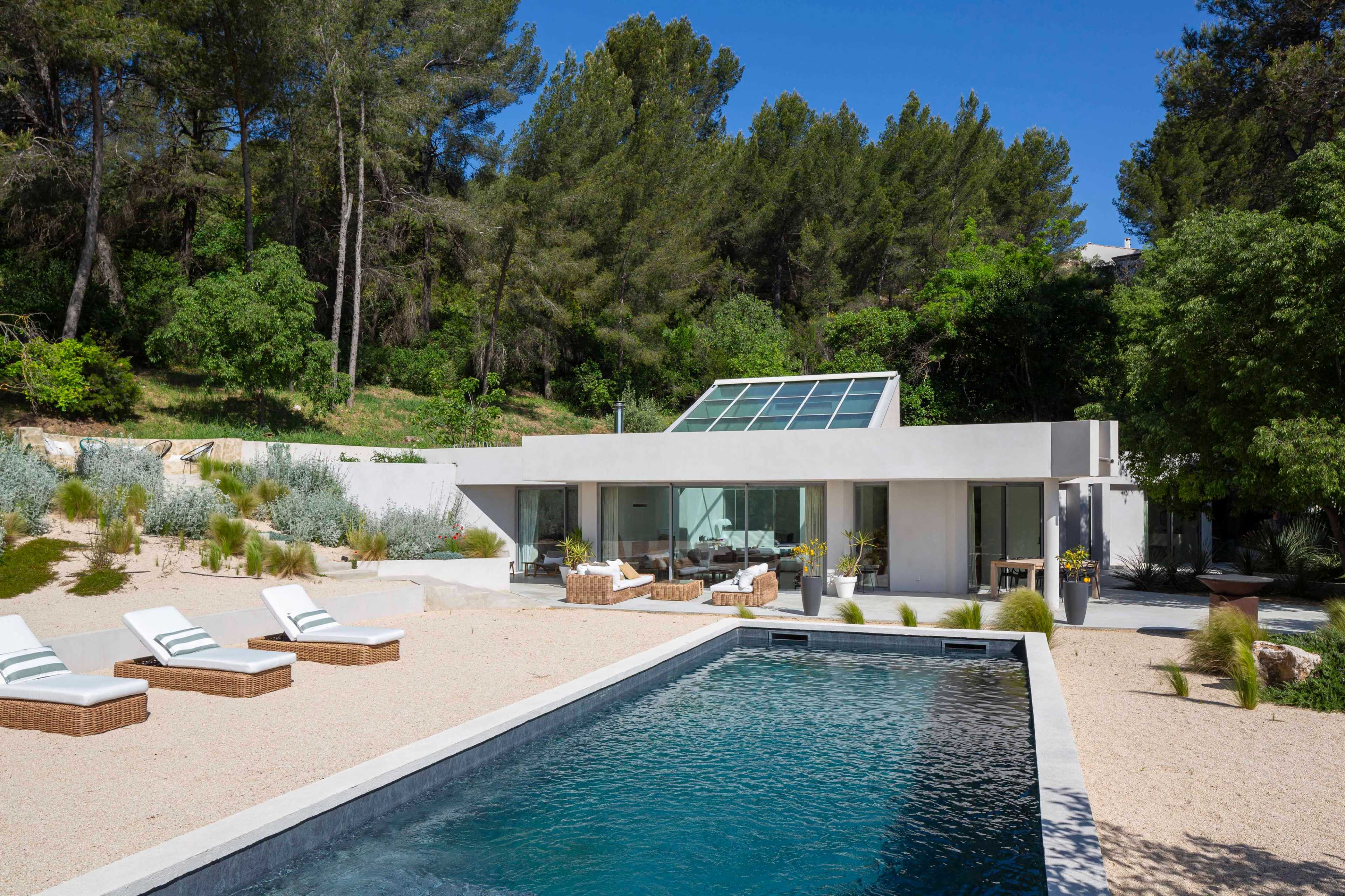 A modern villa with large glass windows overlooks a pool surrounded by lounge chairs and landscaped greenery.