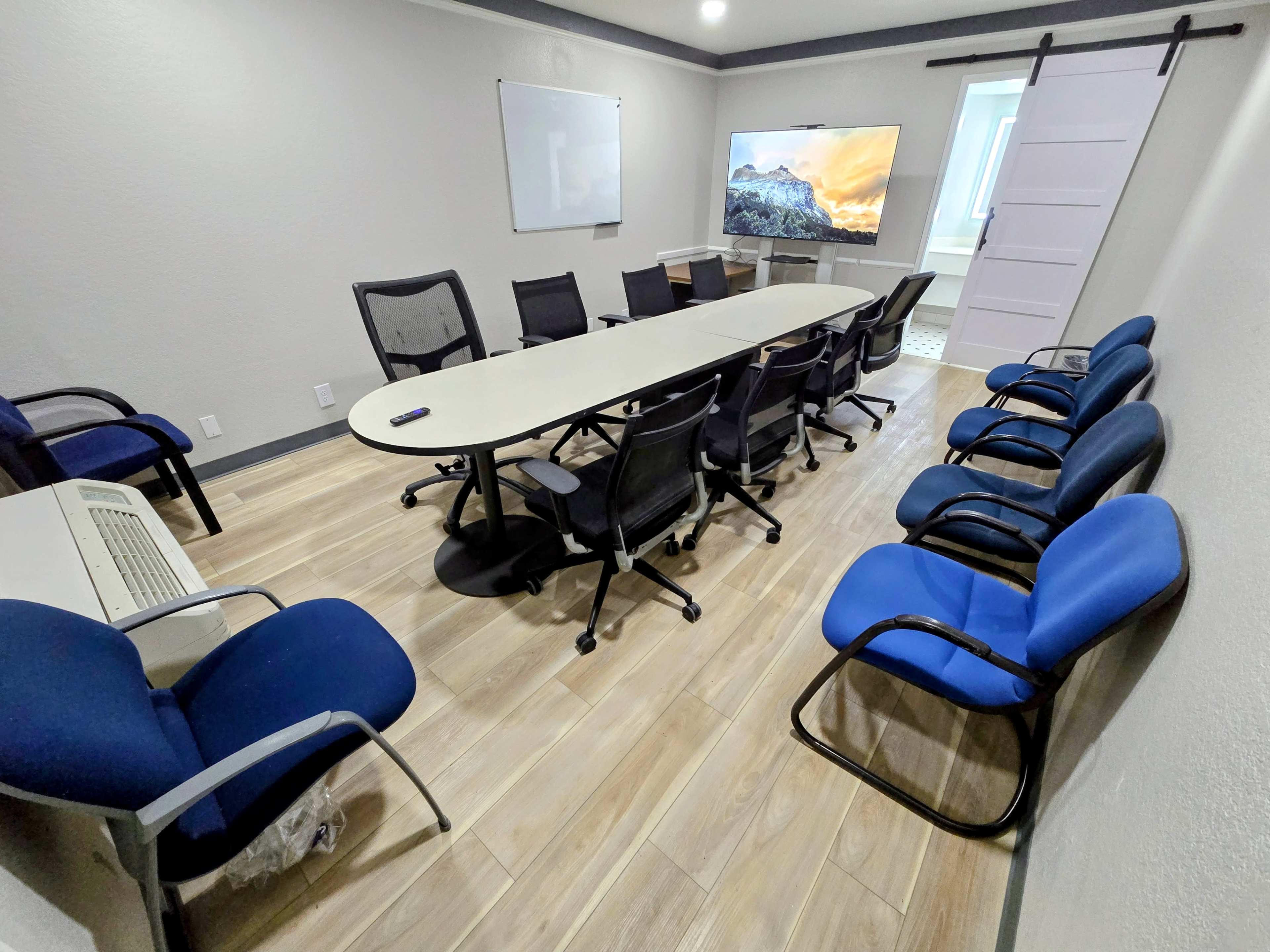 The image shows a modern conference room with a long oval table surrounded by black and blue chairs, a large screen displaying a scenic image, and a whiteboard on the wall.