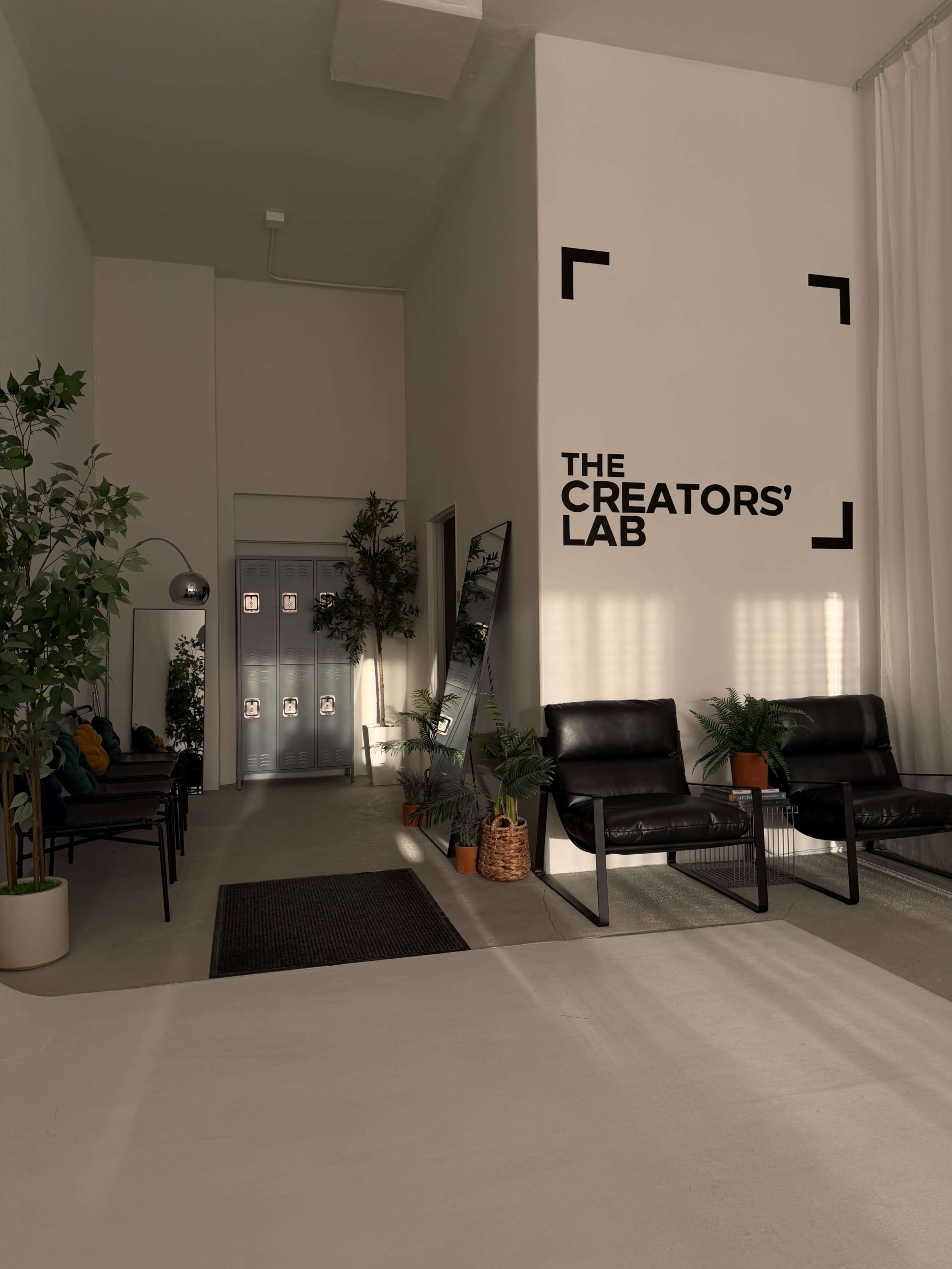 The image shows a minimalist interior of a waiting area with black chairs, plants, and a large sign that reads "THE CREATORS' LAB" on a white wall.