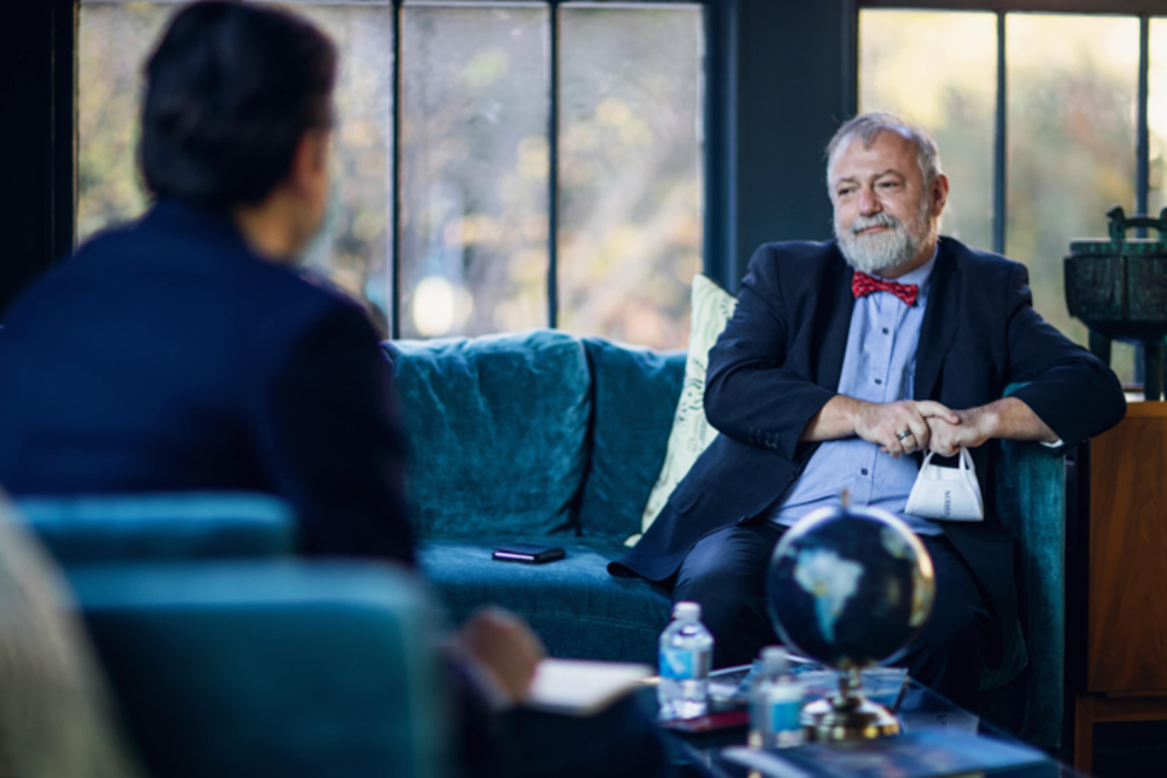 A man in a suit and bow tie sits on a teal sofa during a conversation with another person, while a globe is placed on a table in front of them.