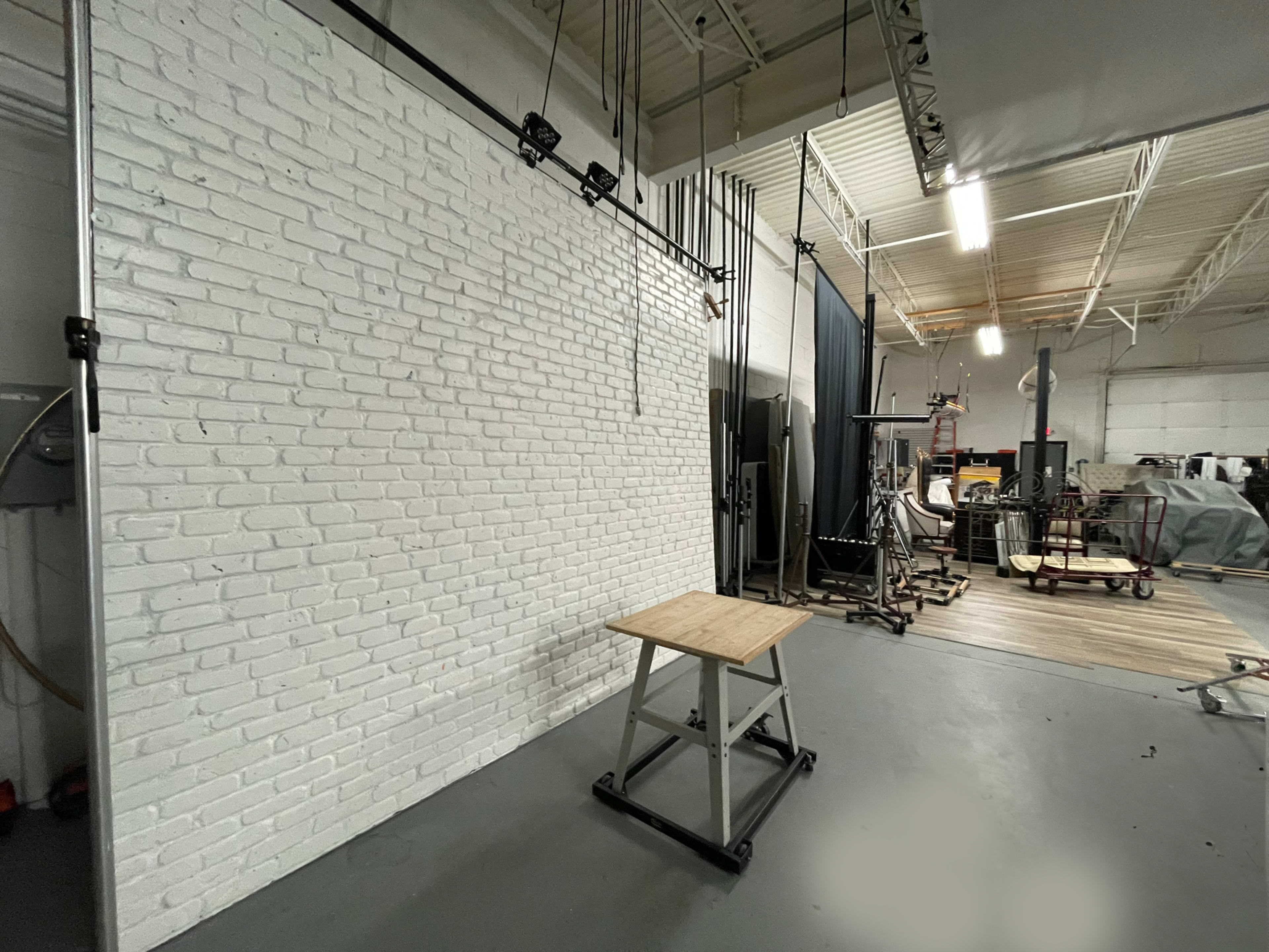 A spacious industrial room with a white brick wall, a wooden table on wheels, and various equipment and backdrops in the background.