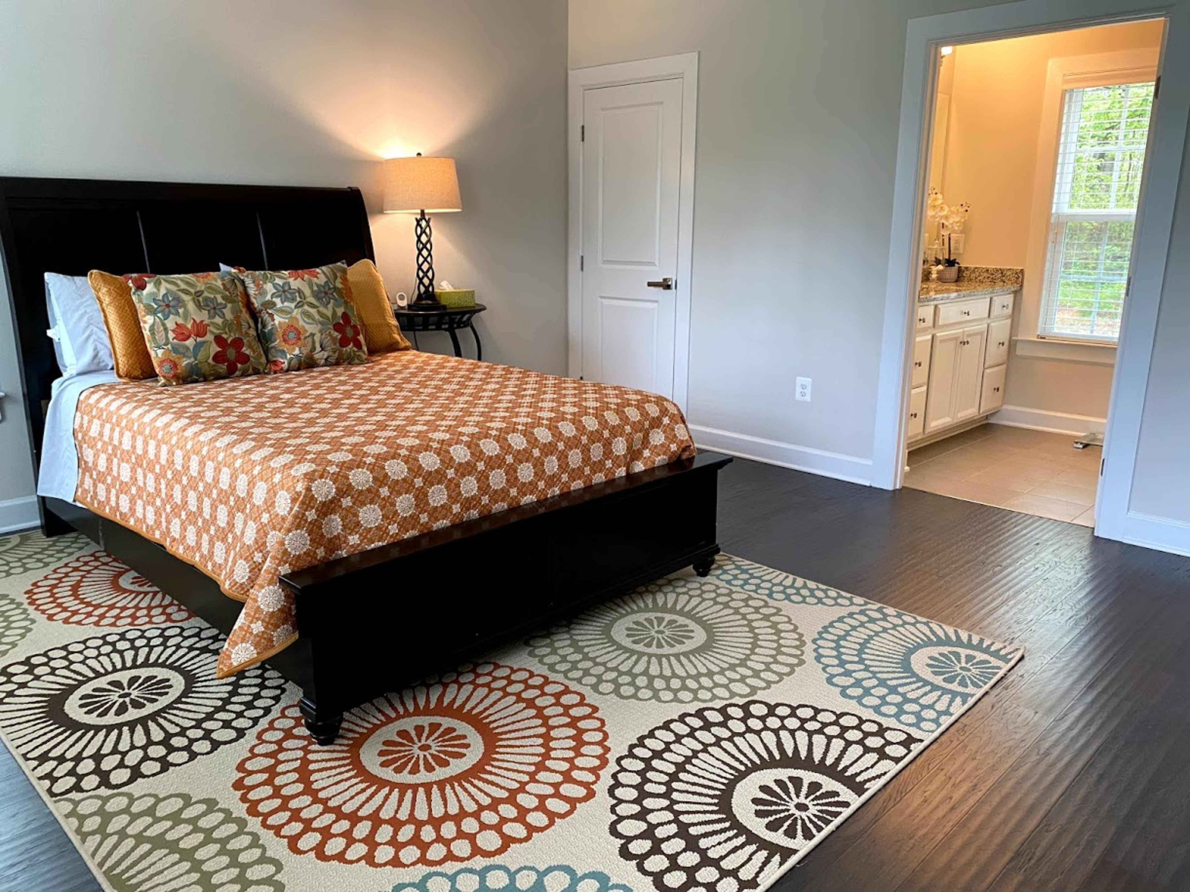 A neatly arranged bedroom with a patterned orange and white bedspread, a wooden bed frame, and an adjacent bathroom visible through an open door.
