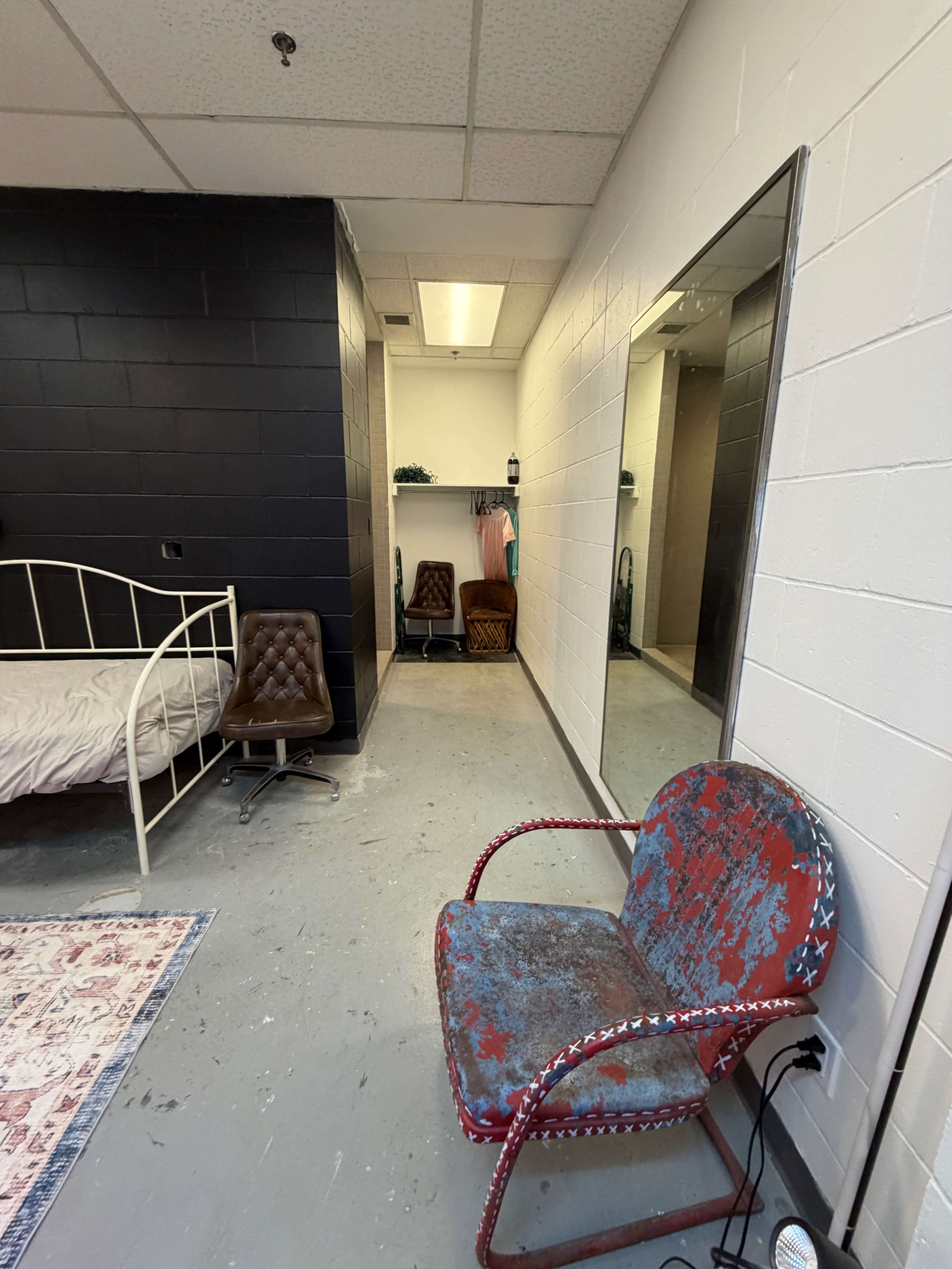 The image shows a sparsely decorated room with a bed, a large mirror, and a worn chair, featuring black and white walls.