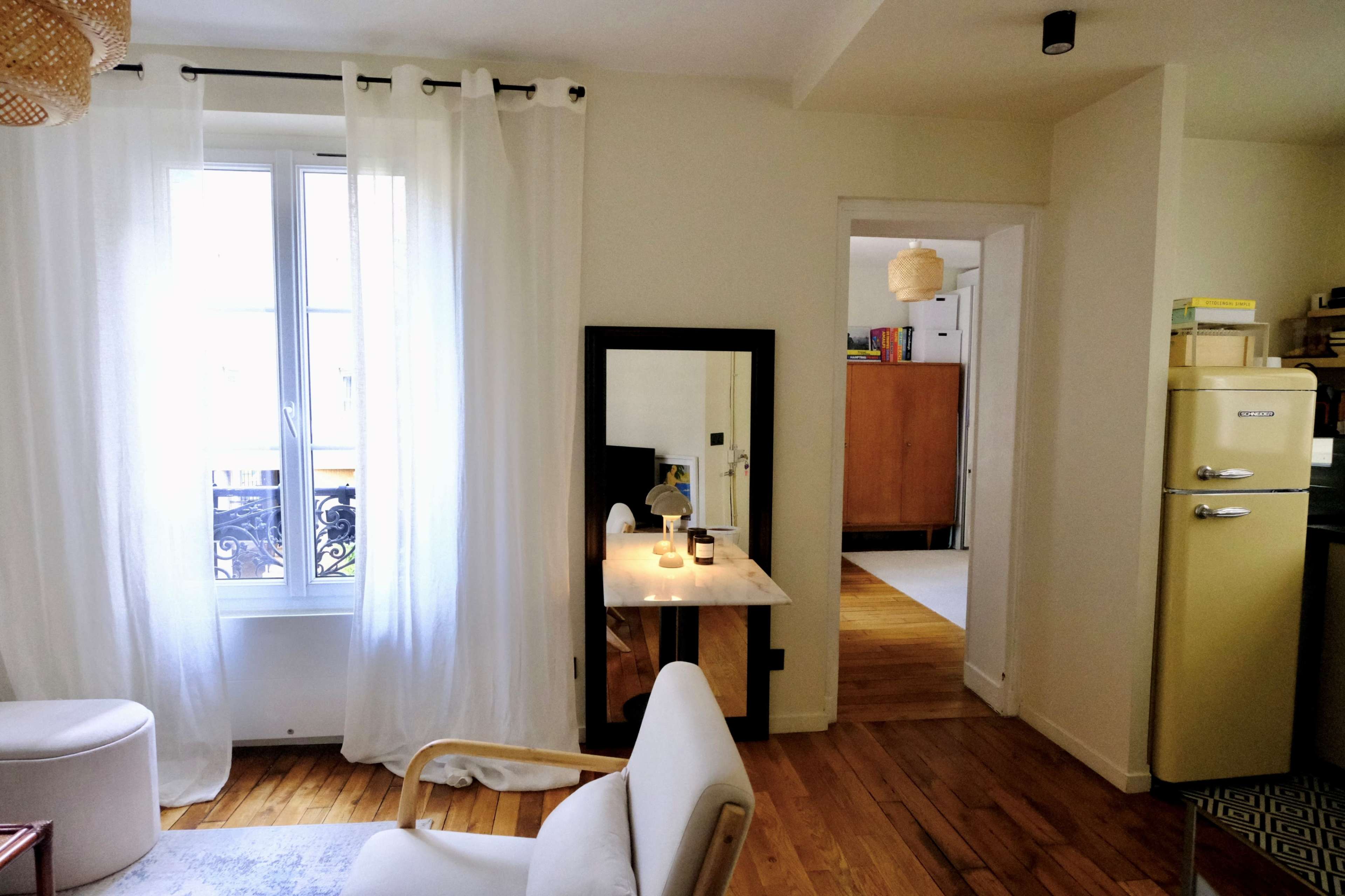 The image shows a bright, modern living space featuring a white curtain-draped window, a small marble table with candles, and an open doorway leading to another room.