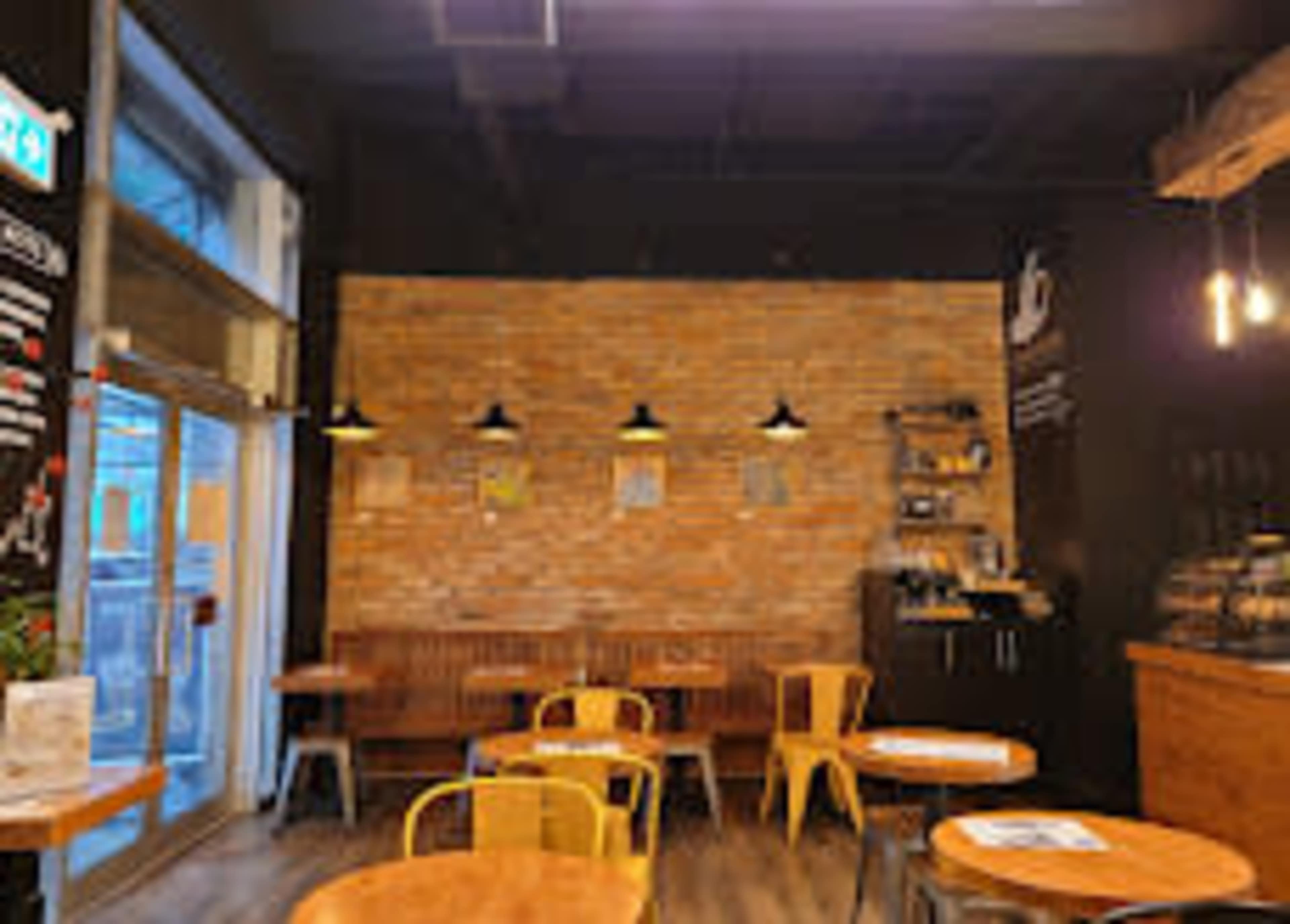 The image shows an interior of a café featuring brick walls, wooden benches, round tables, and hanging light fixtures.