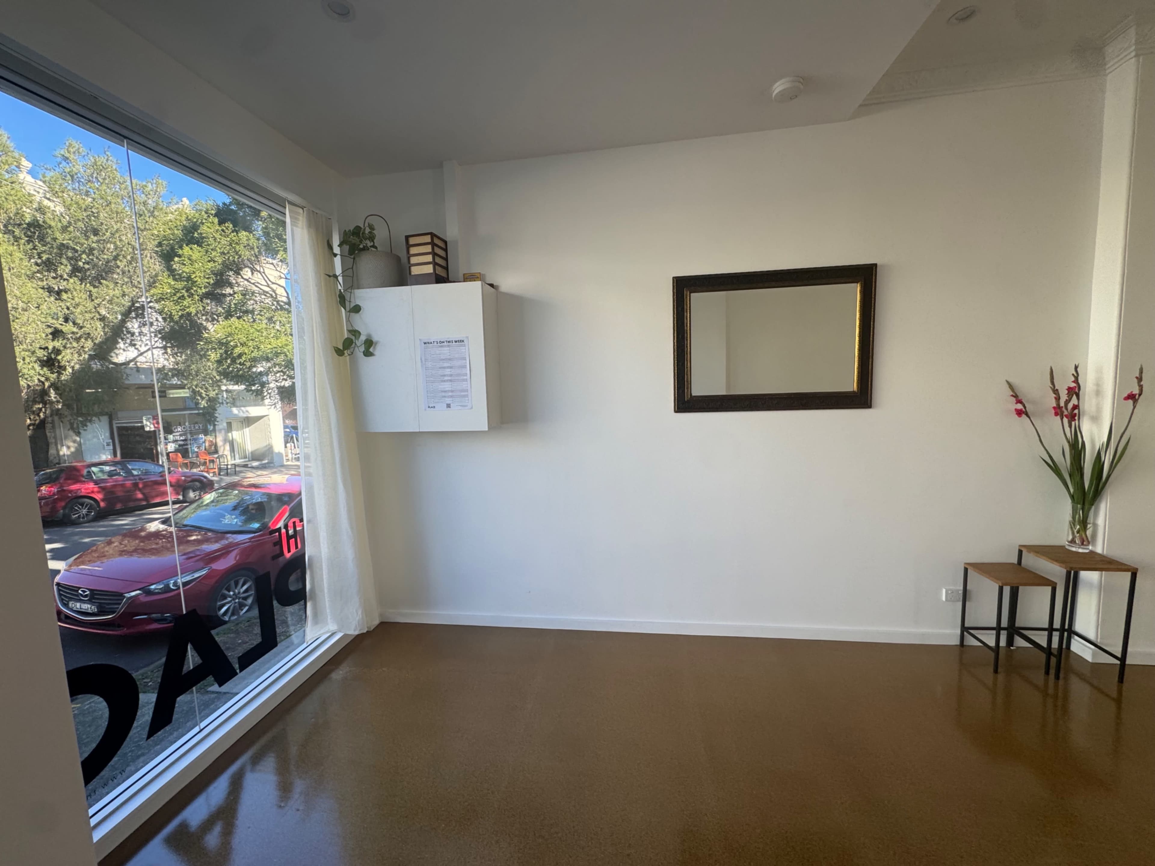 The image shows a minimalist interior space with a large window, a mirror on the wall, a small cabinet, and two wooden stools.