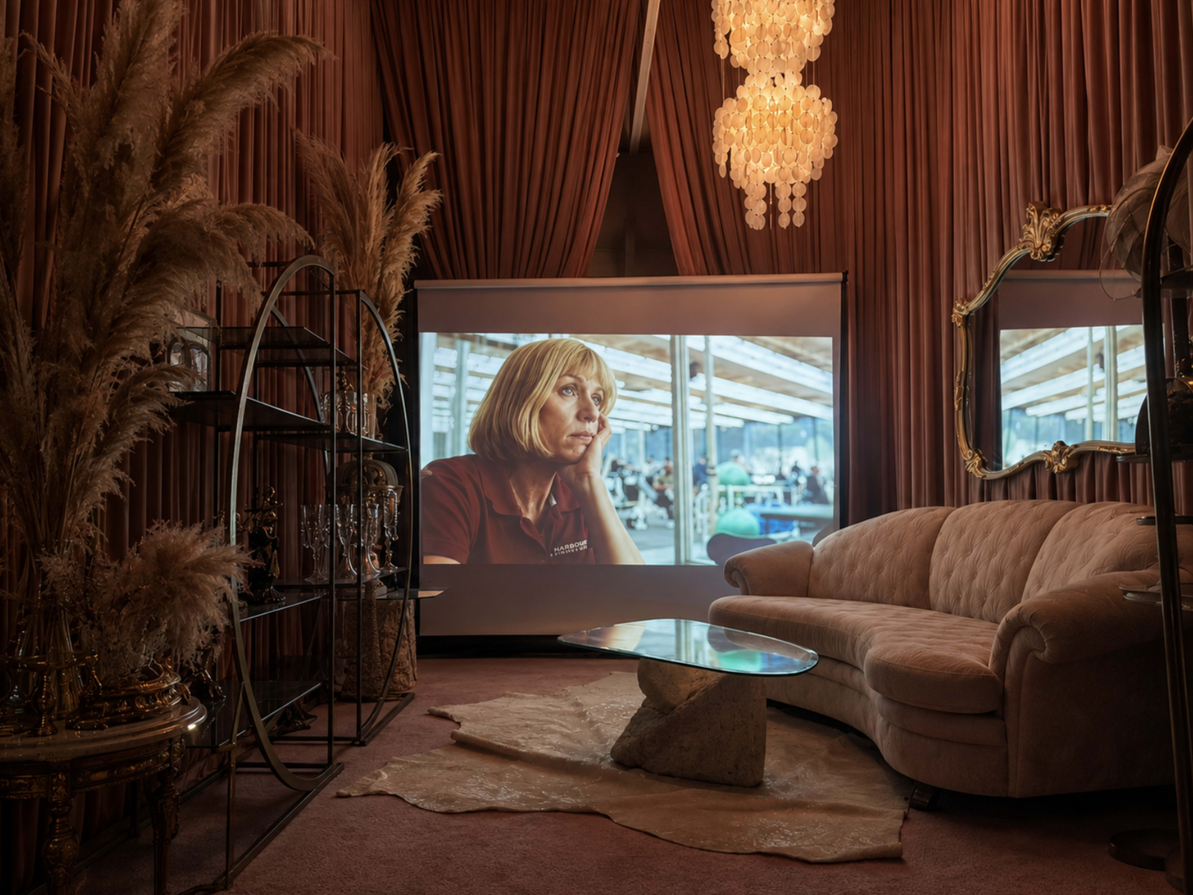 The image shows a cozy indoor space with a beige couch, a stylish coffee table, and a large screen displaying a woman in deep thought, surrounded by decorative plants and elegant lighting.